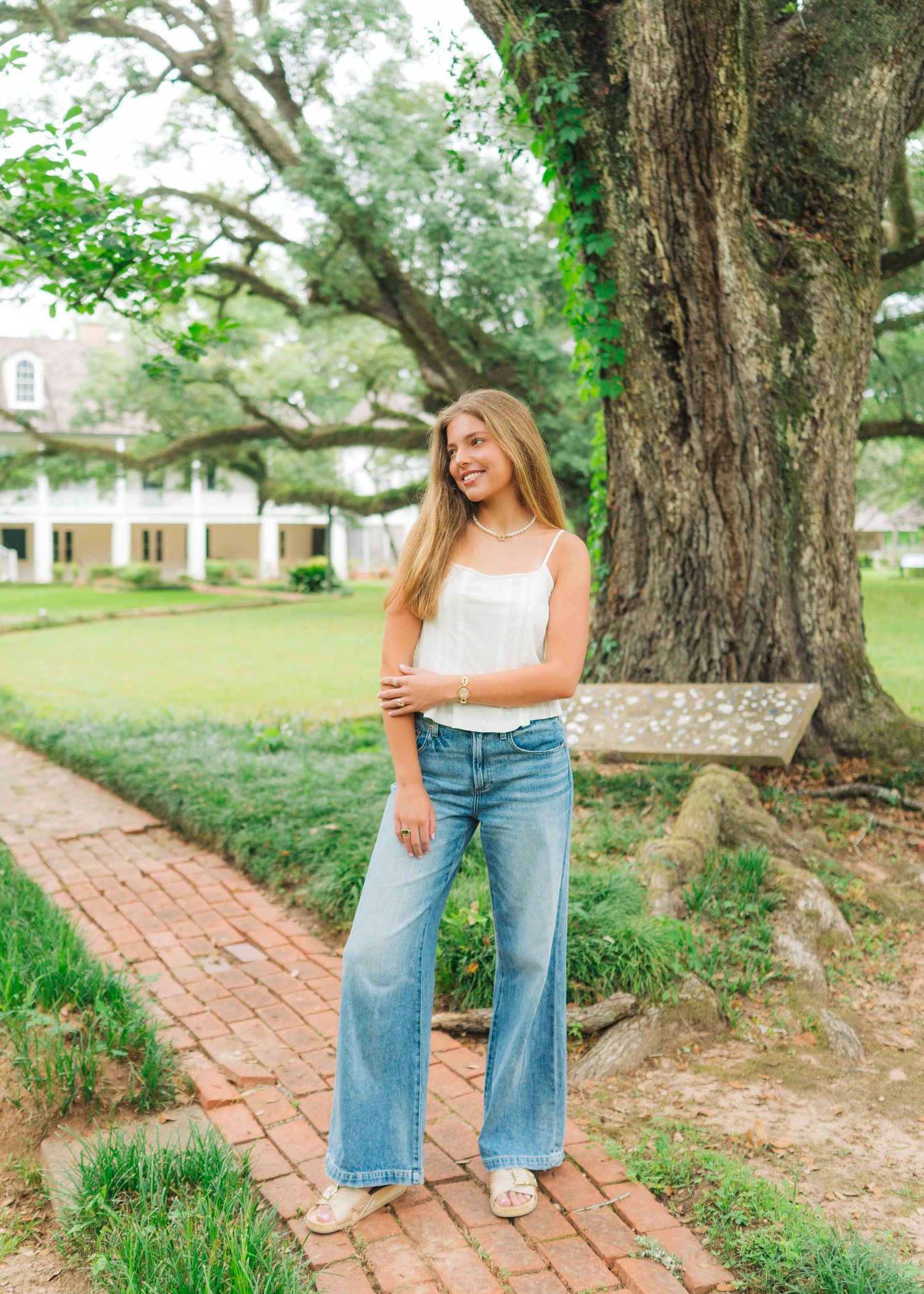 Smiling teen girl in wide-leg jeans and white cami stands on brick path near historic oak.