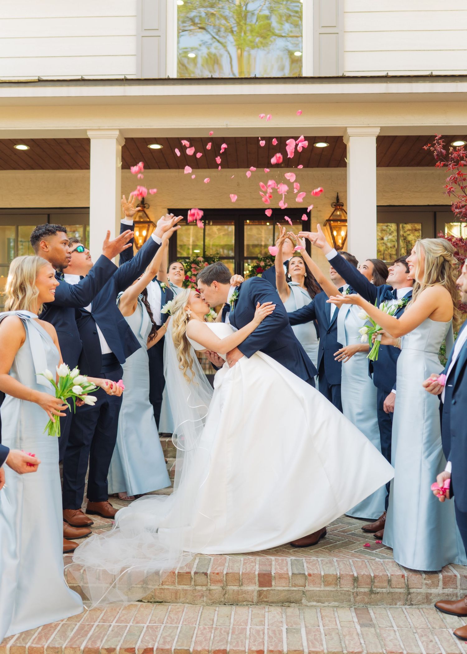 Wedding party tossing pink rose petals over kissing bride and groom on brick steps outdoors.