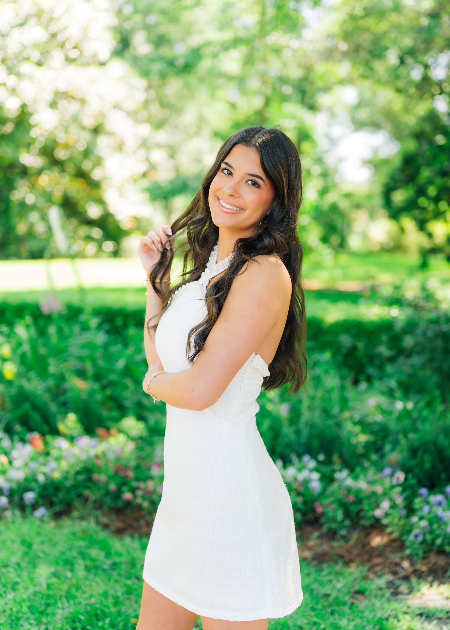 Young woman in white dress smiling in lush garden with colorful flowers and green trees.
