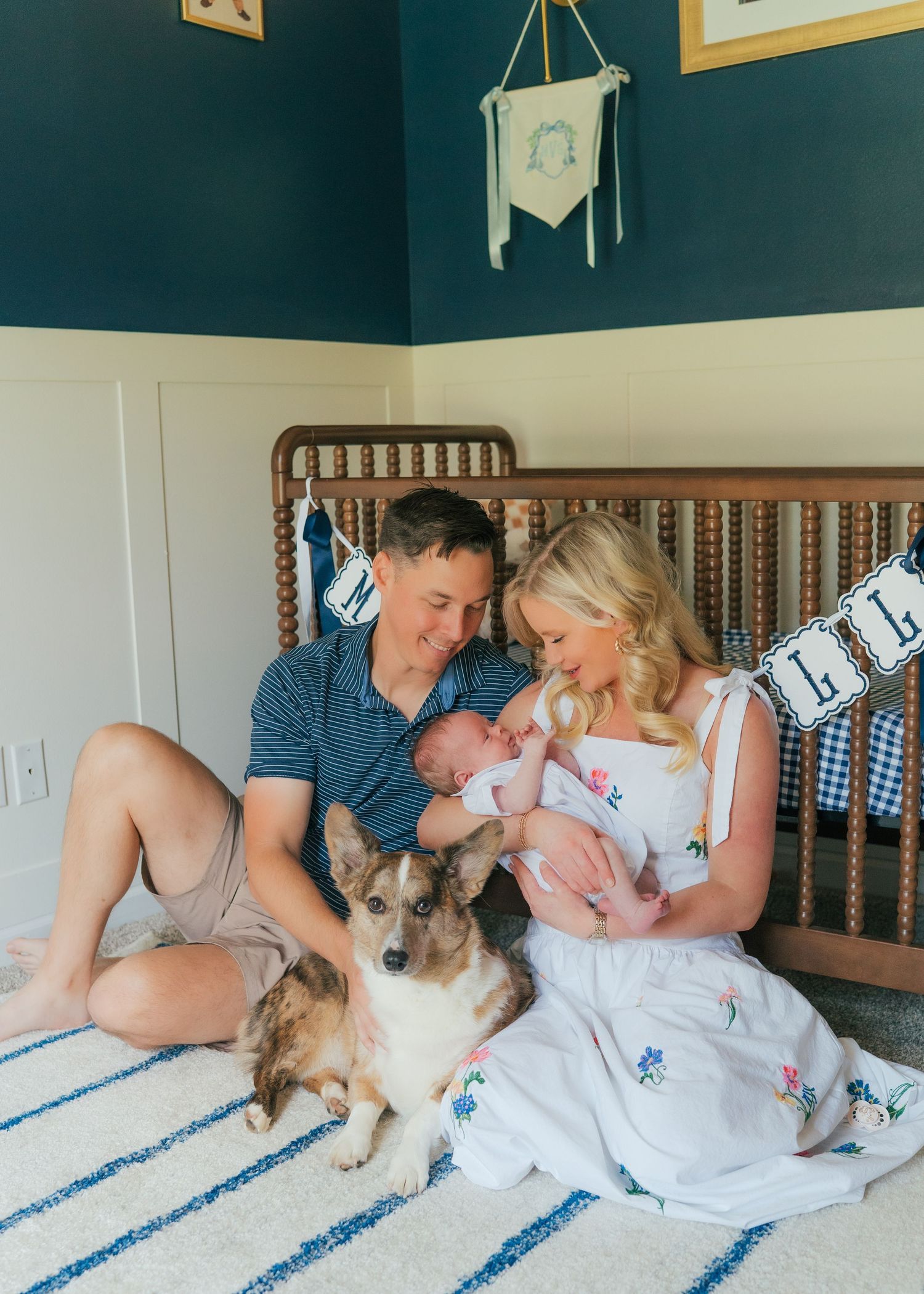 A family with a newborn baby and corgi dog sit together in a navy blue nursery room.