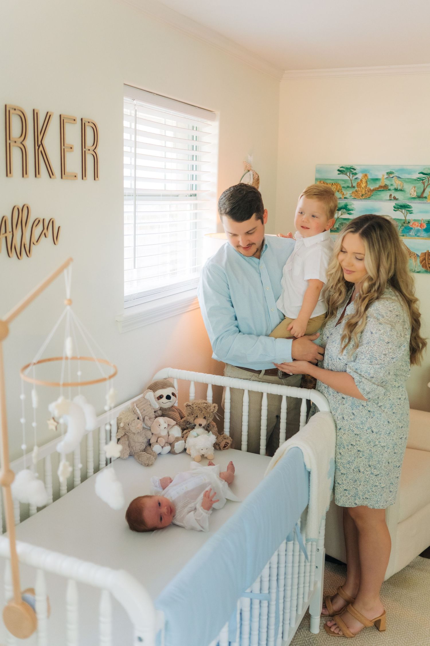 Happy family of four gazes at newborn lying in white crib in a decorated nursery with name on wall.