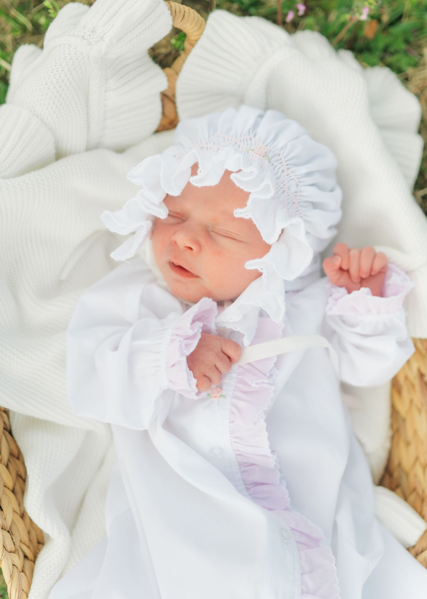 Sleeping newborn baby in white smocked gown and ruffled bonnet nestled in a wicker basket with white blanket.