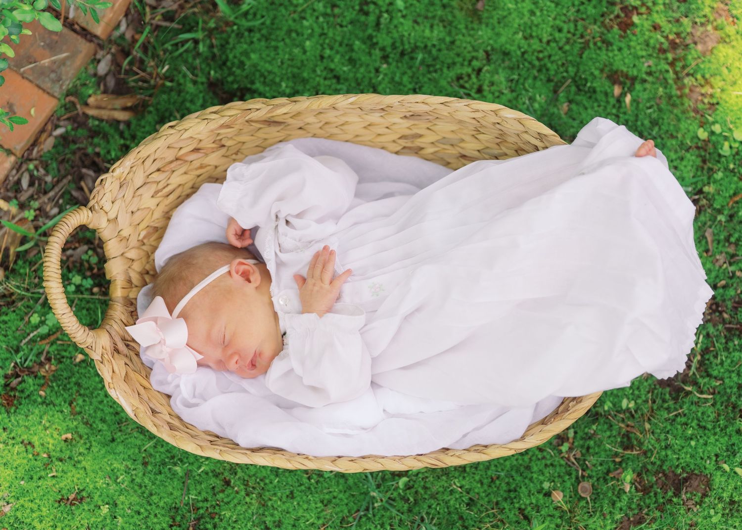 Newborn baby sleeping peacefully in a wicker basket wrapped in white cloth on green moss outdoors.