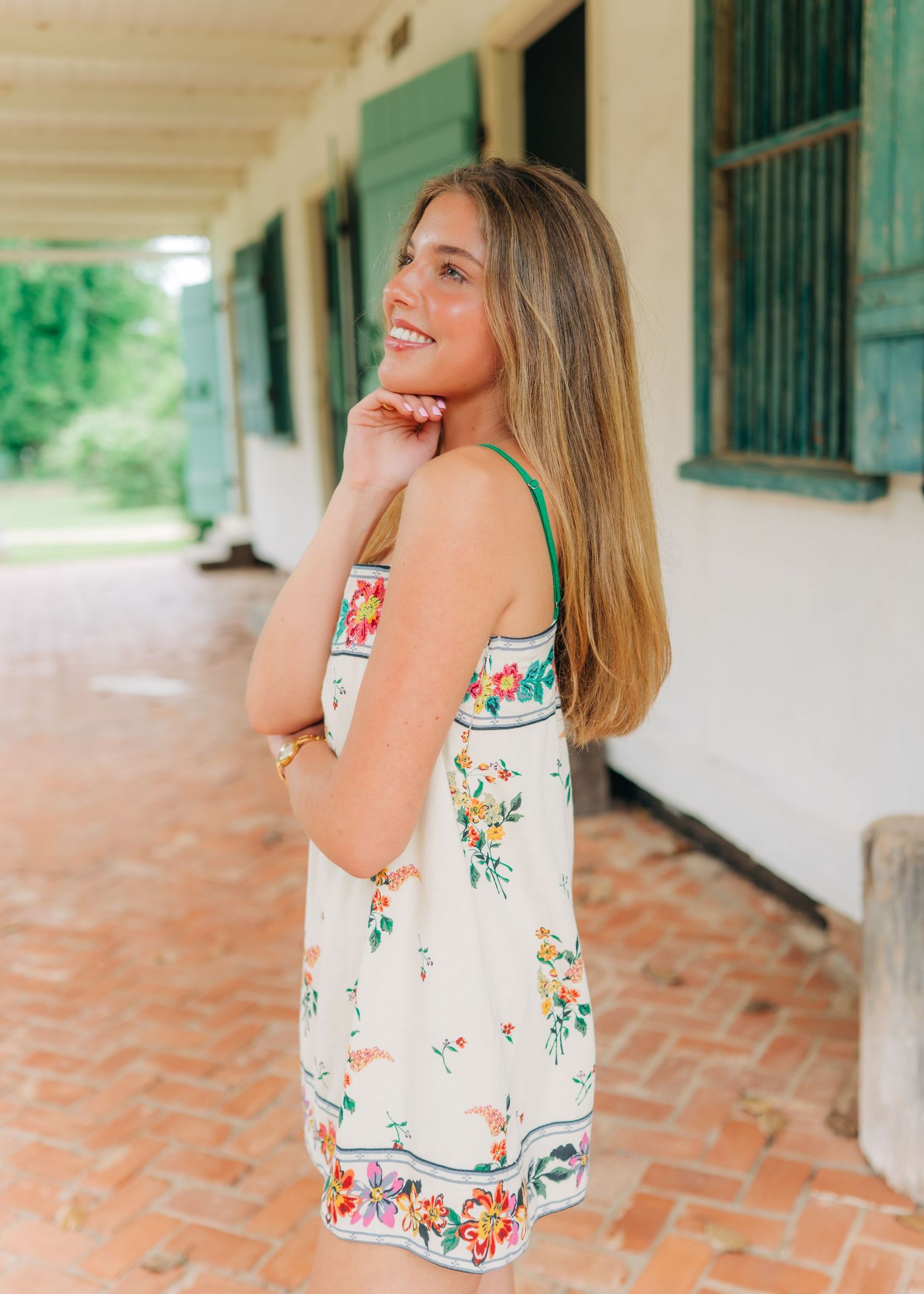Smiling young woman in white floral sundress with green straps posing on brick porch.