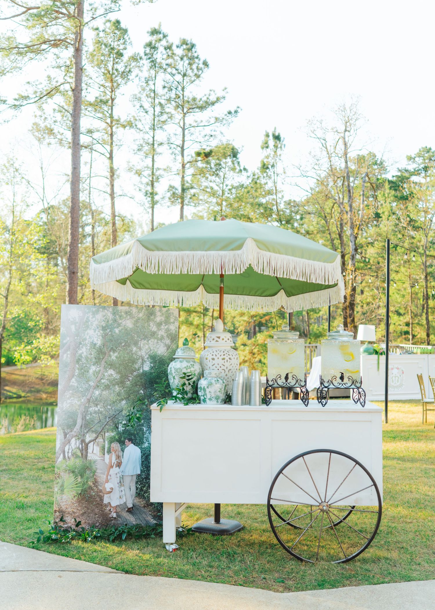 White beverage cart with green fringed umbrella and floral arrangements at outdoor wedding.