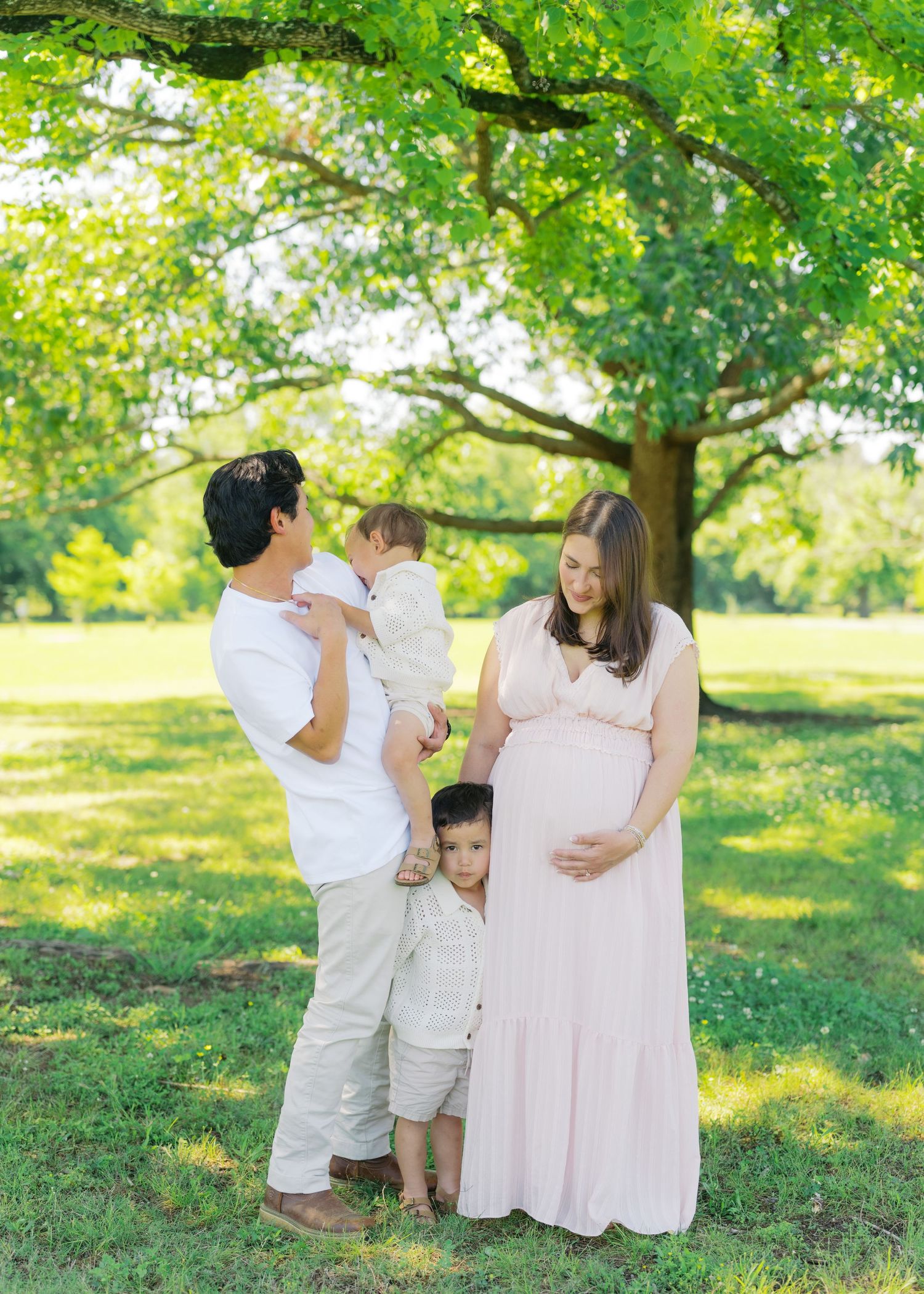 Pregnant woman in pink dress with husband and two young children posing outdoors under a large green tree.
