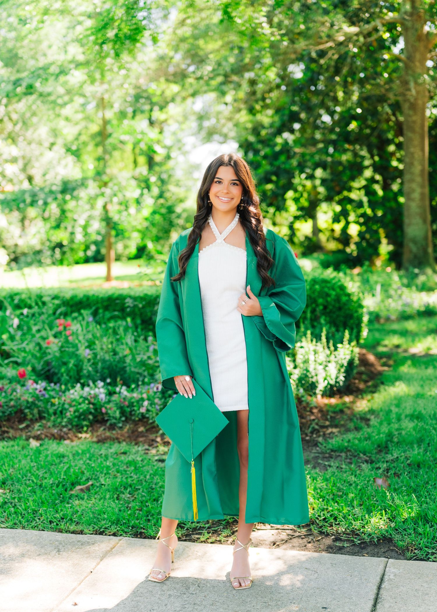 Smiling graduate in green cap and gown over white dress poses outdoors in lush garden setting.