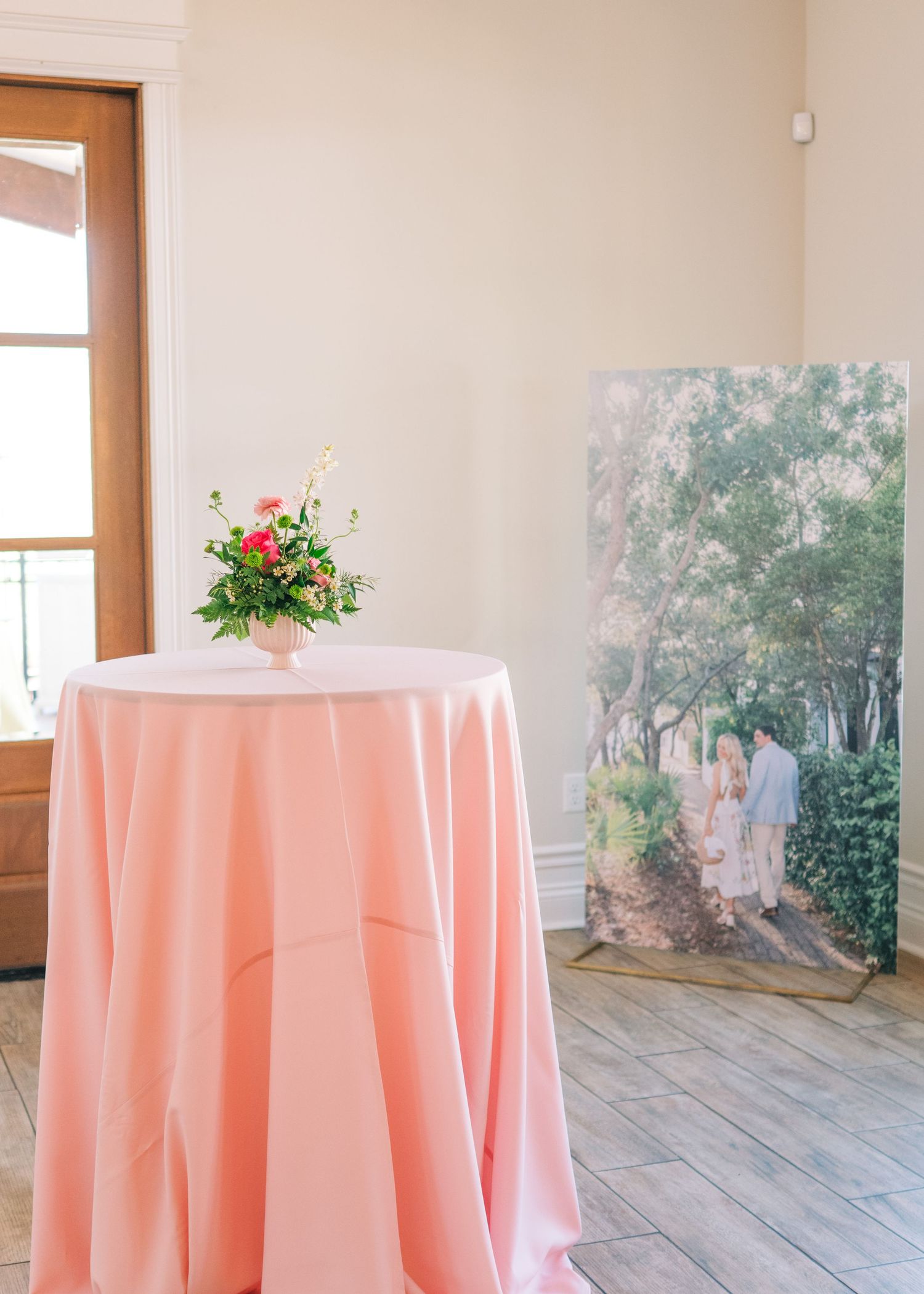 Pink tablecloth cocktail table with floral arrangement beside large engagement photo display.