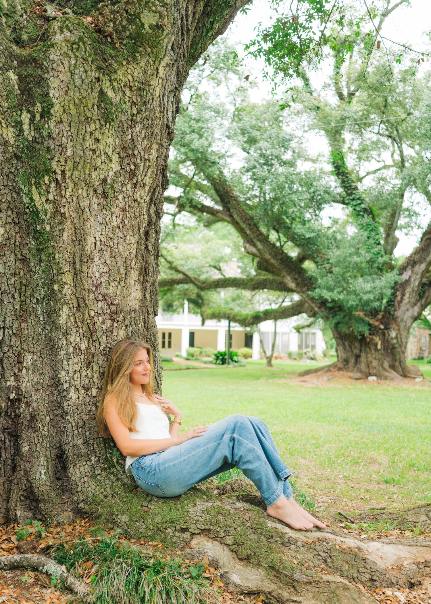 Woman in white top and jeans sitting against large oak tree roots on green lawn.
