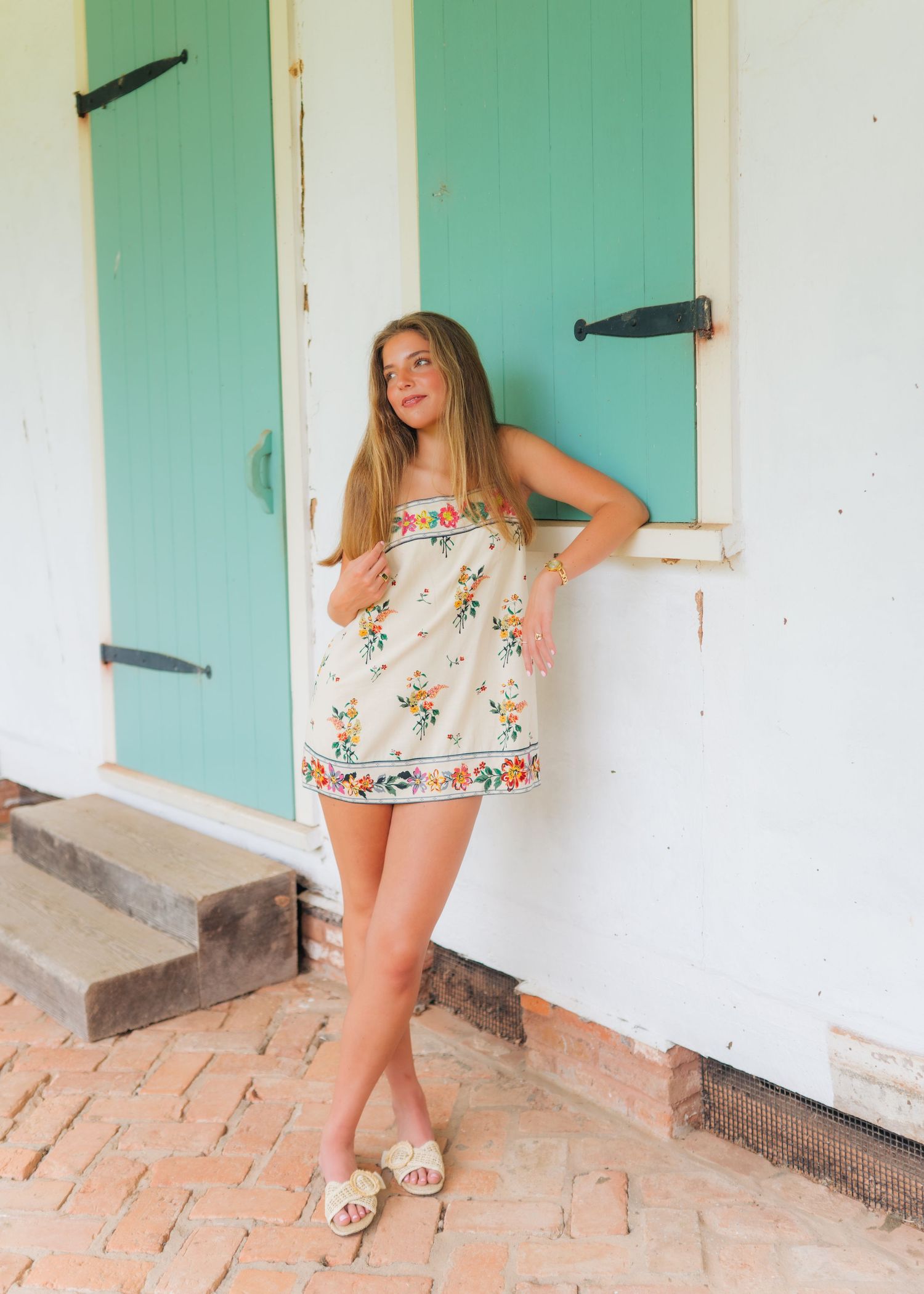 Young woman in a floral embroidered mini dress poses by turquoise barn doors on a brick patio.