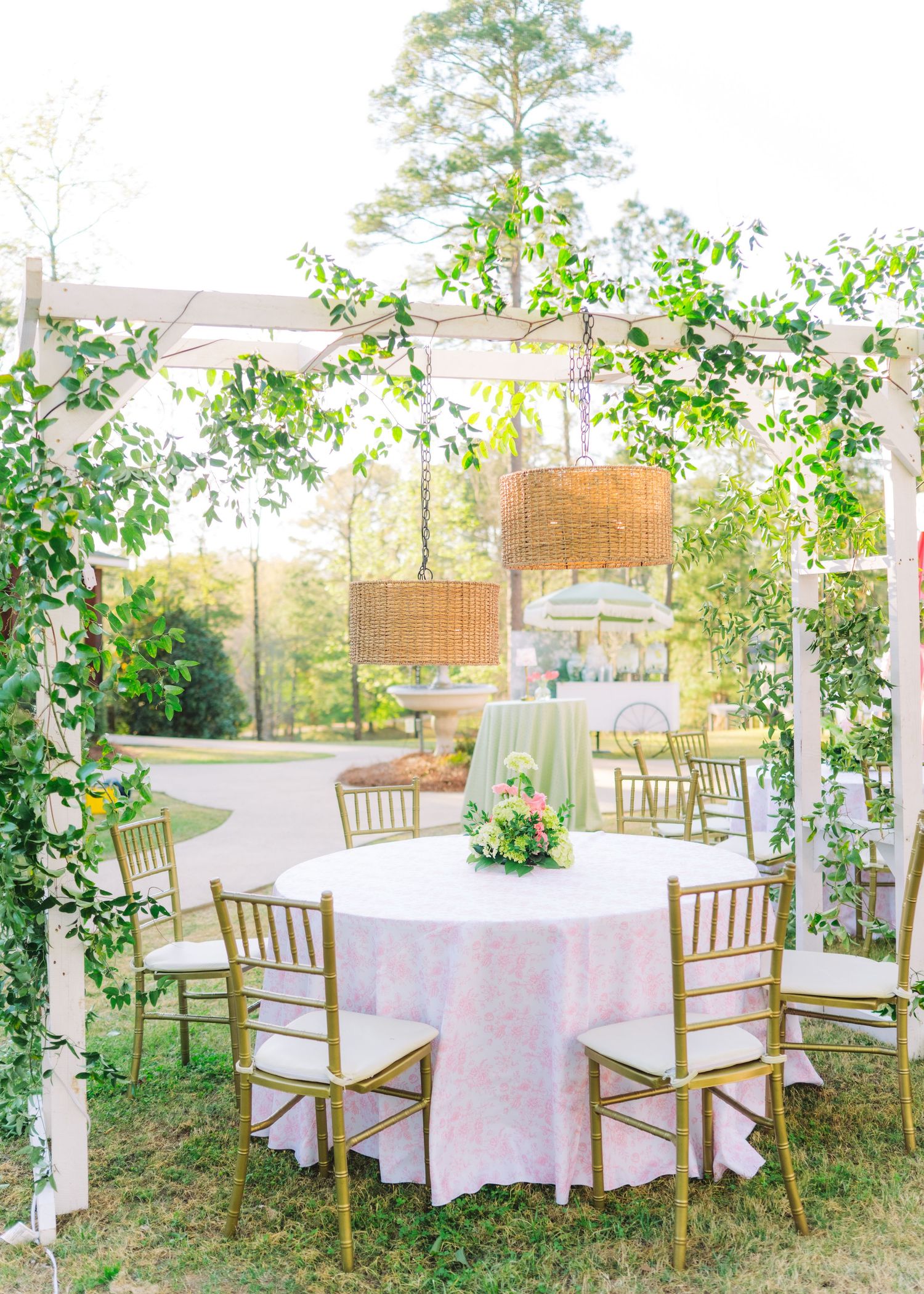 Outdoor wedding reception table with pink linen under white pergola draped with lush greenery and wicker chandeliers.