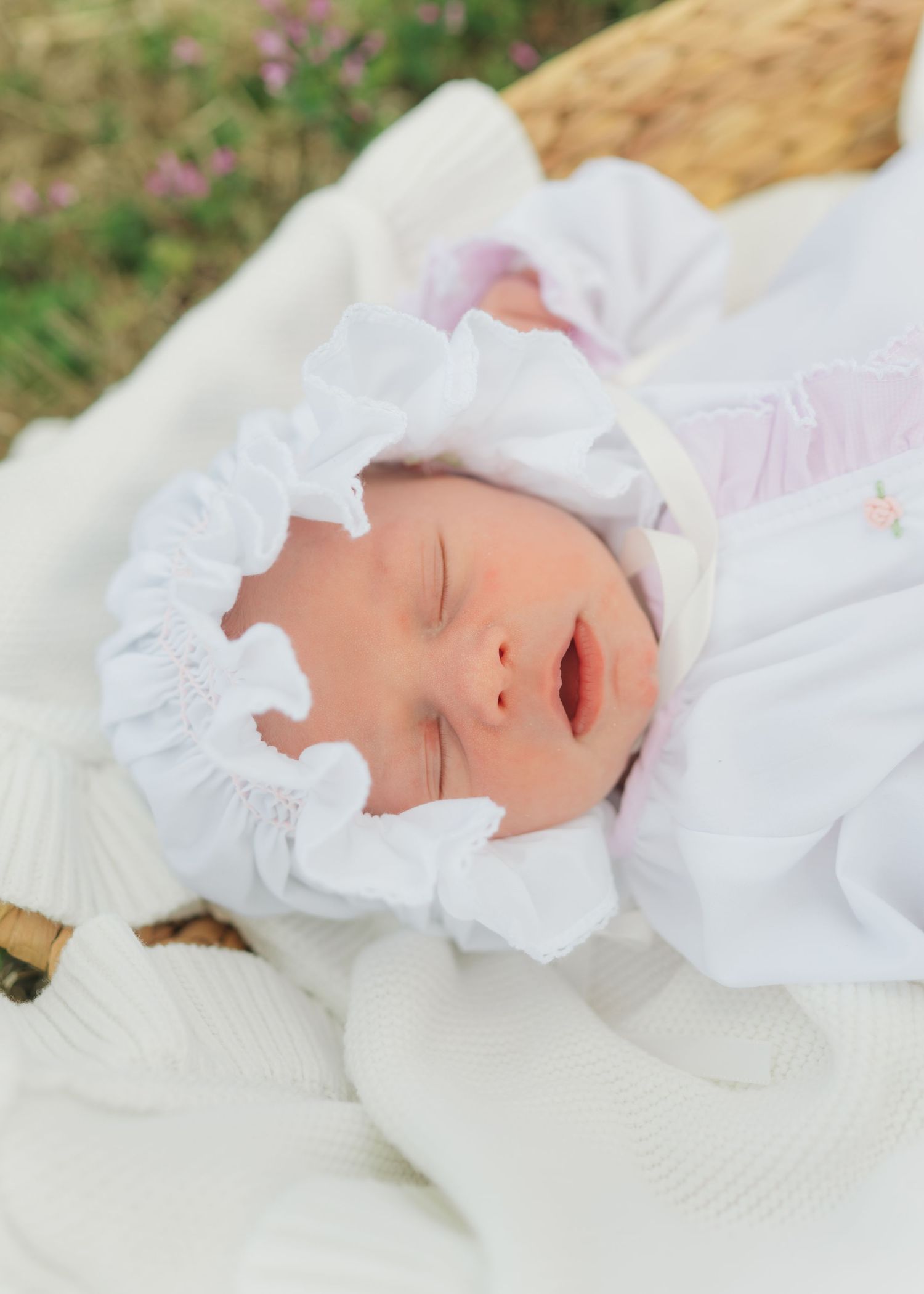 Newborn baby sleeping peacefully in a white ruffled bonnet and outfit, nestled in a wicker basket outdoors.