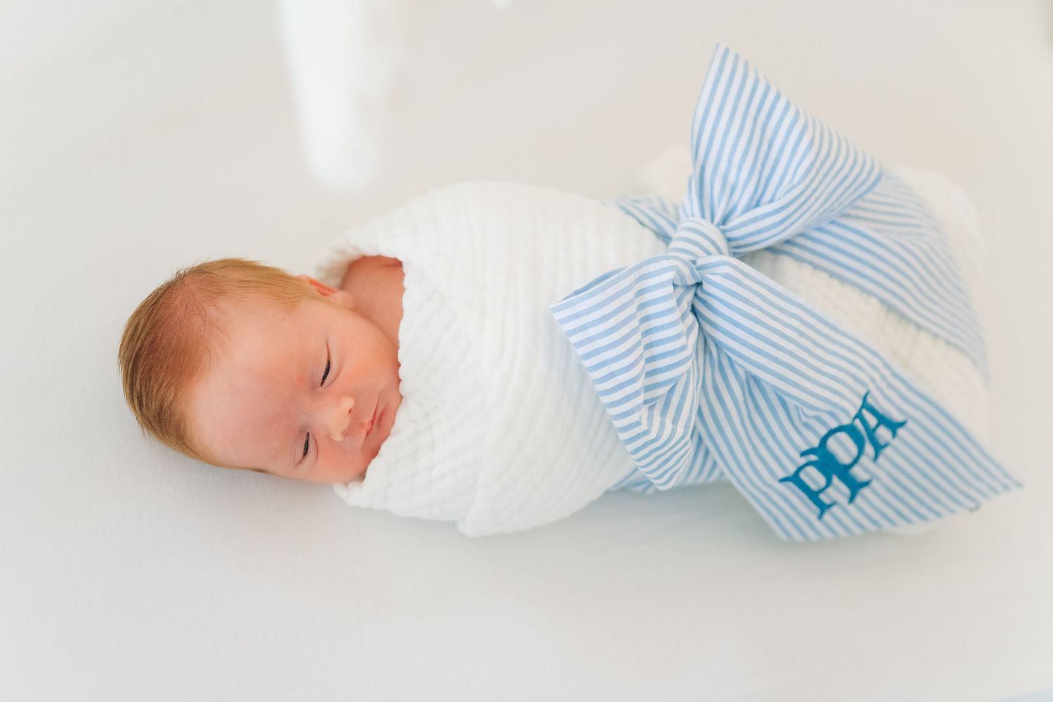 Sleeping newborn baby swaddled in white wrap with a blue seersucker bow monogrammed with PPH.