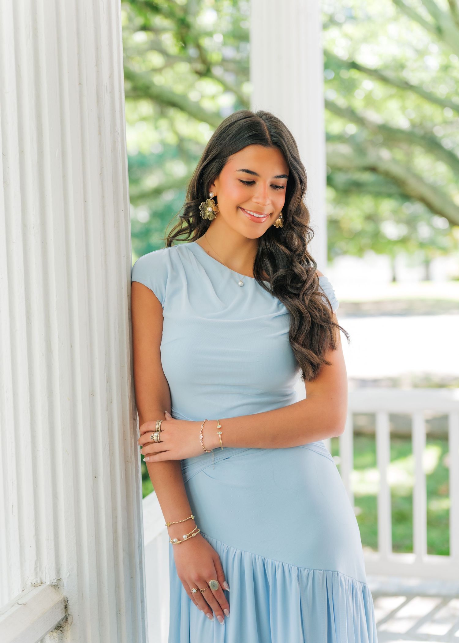 Young woman in light blue midi dress leans against white column, smiling downward, wearing gold earrings and bracelets.