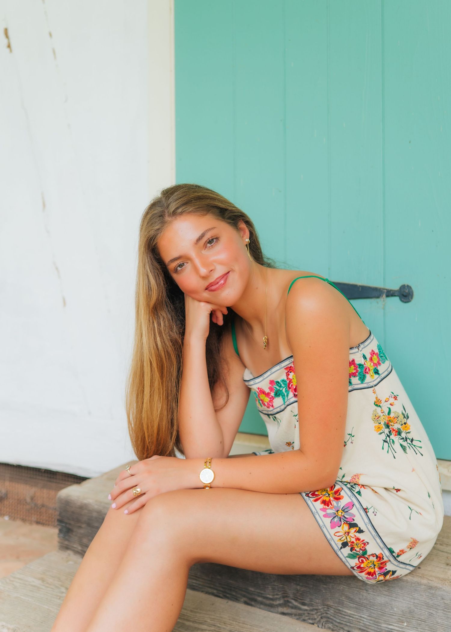 Young woman in floral sundress sitting on steps by turquoise door, resting chin on hand, smiling.