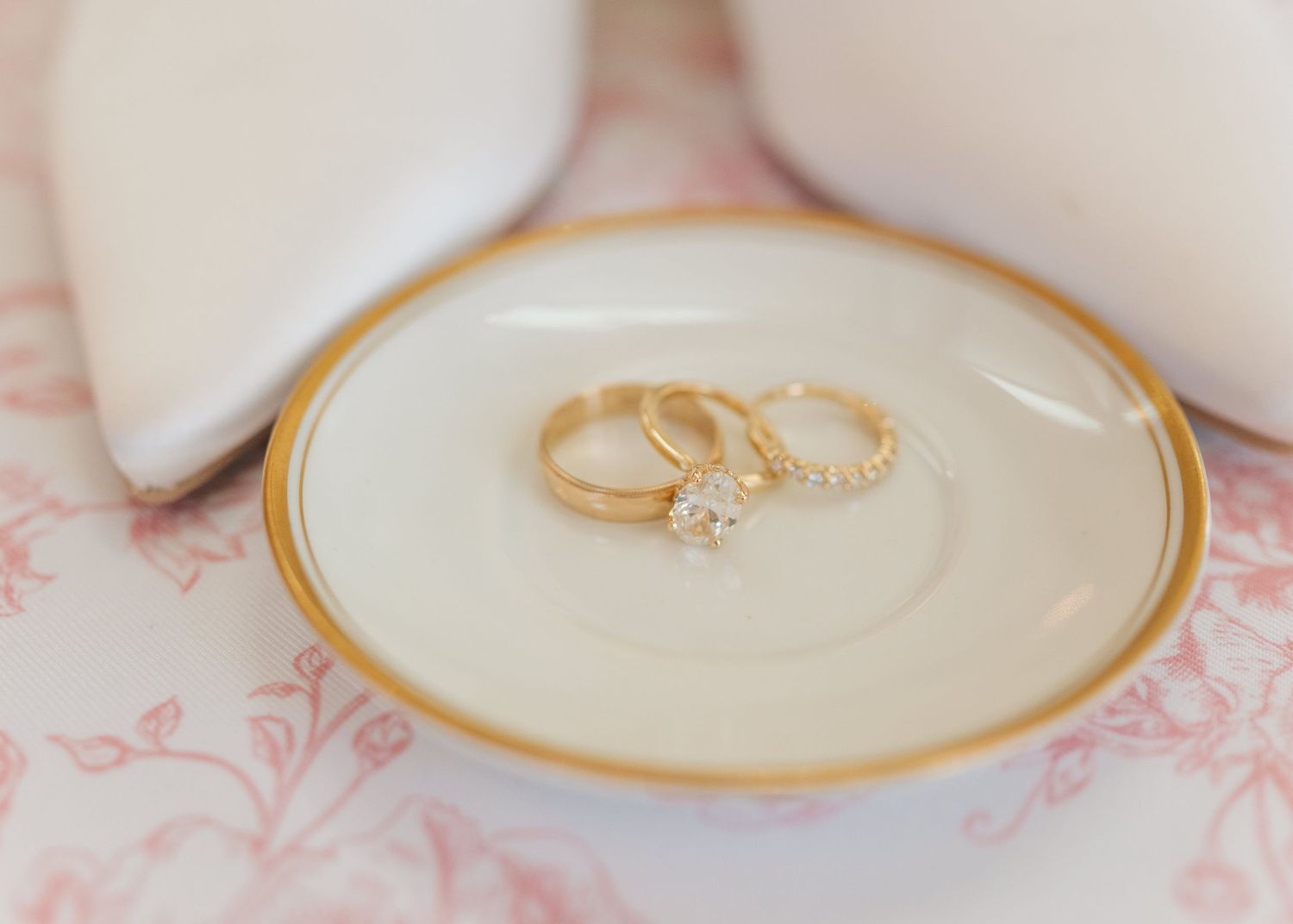 Three gold wedding rings with diamonds displayed on a small white and gold decorative plate near white bridal shoes.
