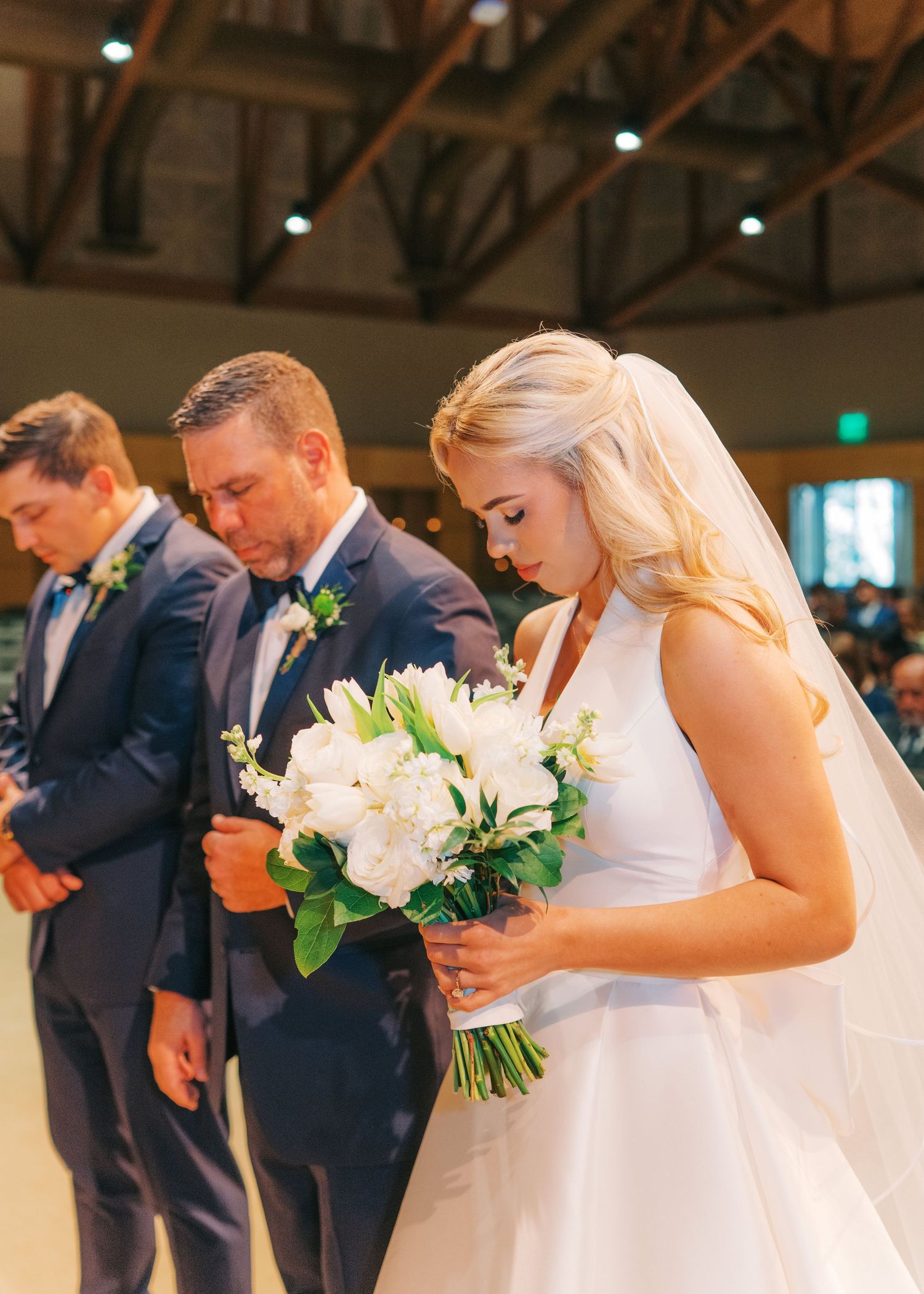Bride in white gown holds white bouquet with groom in navy suit bowing heads in prayer during wedding ceremony.