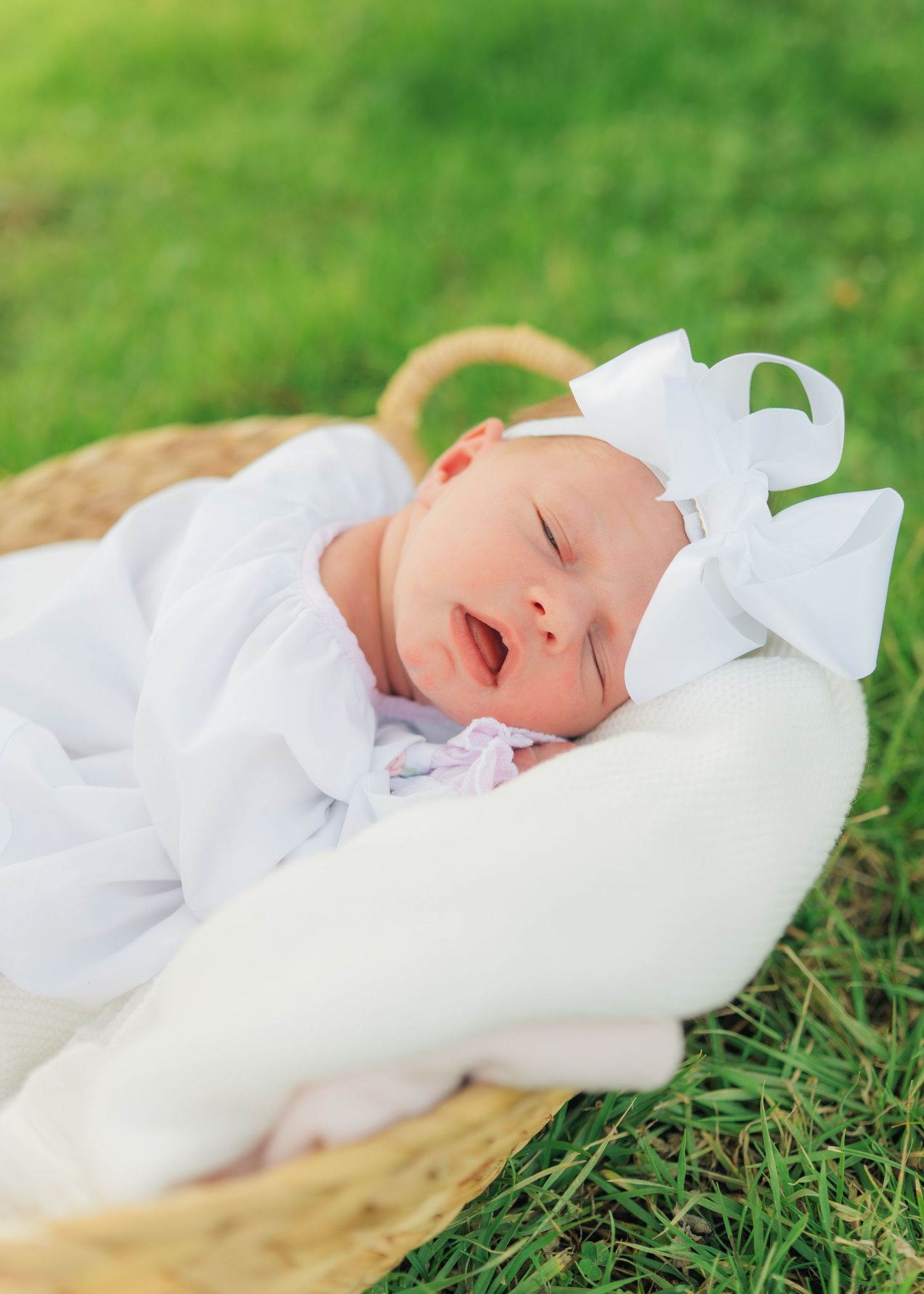 Sleeping newborn baby in white outfit and bow headband nestled in a wicker basket on green grass.