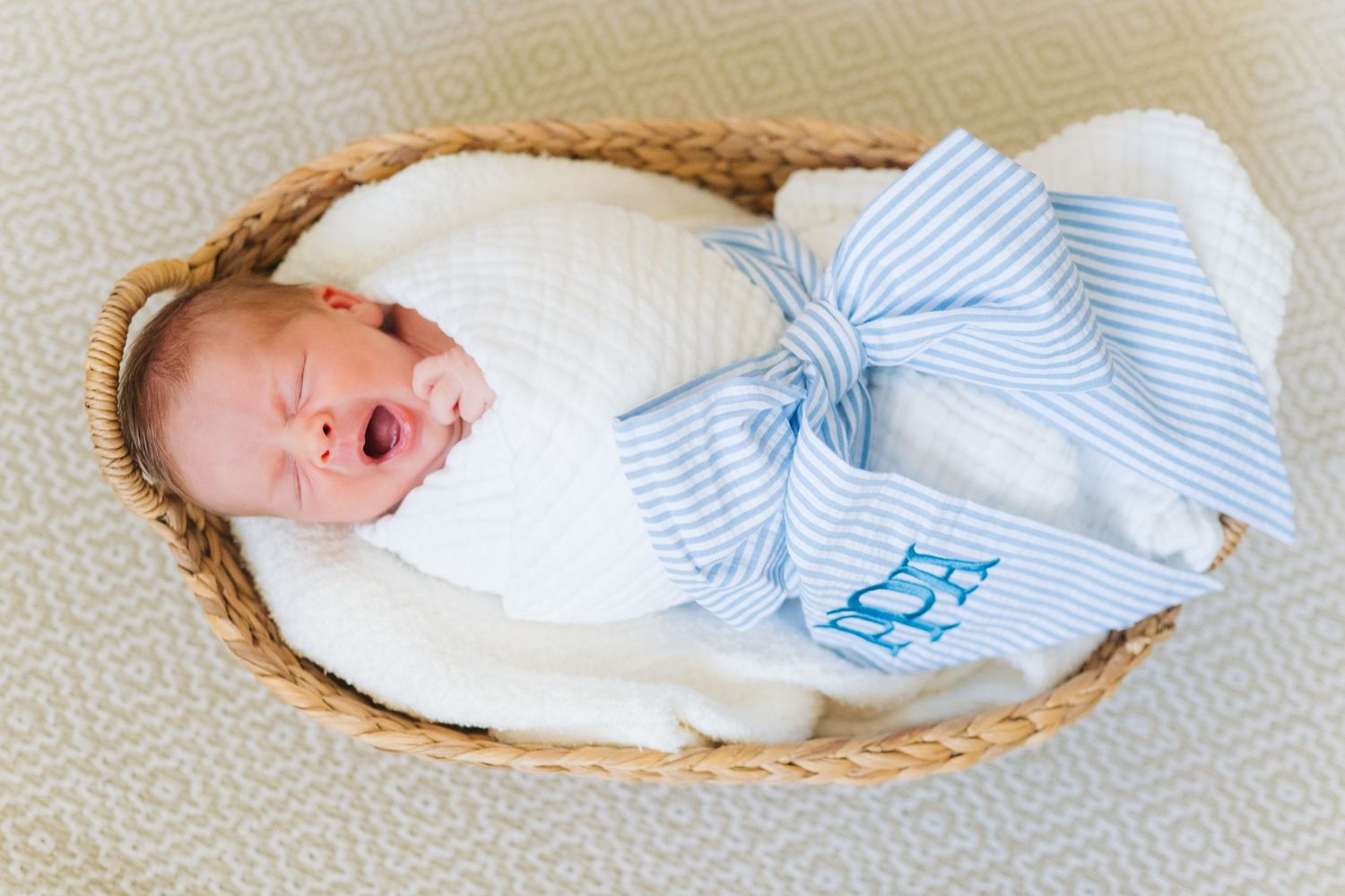 Newborn baby swaddled in white blanket with blue monogrammed bow, sleeping in a wicker basket.
