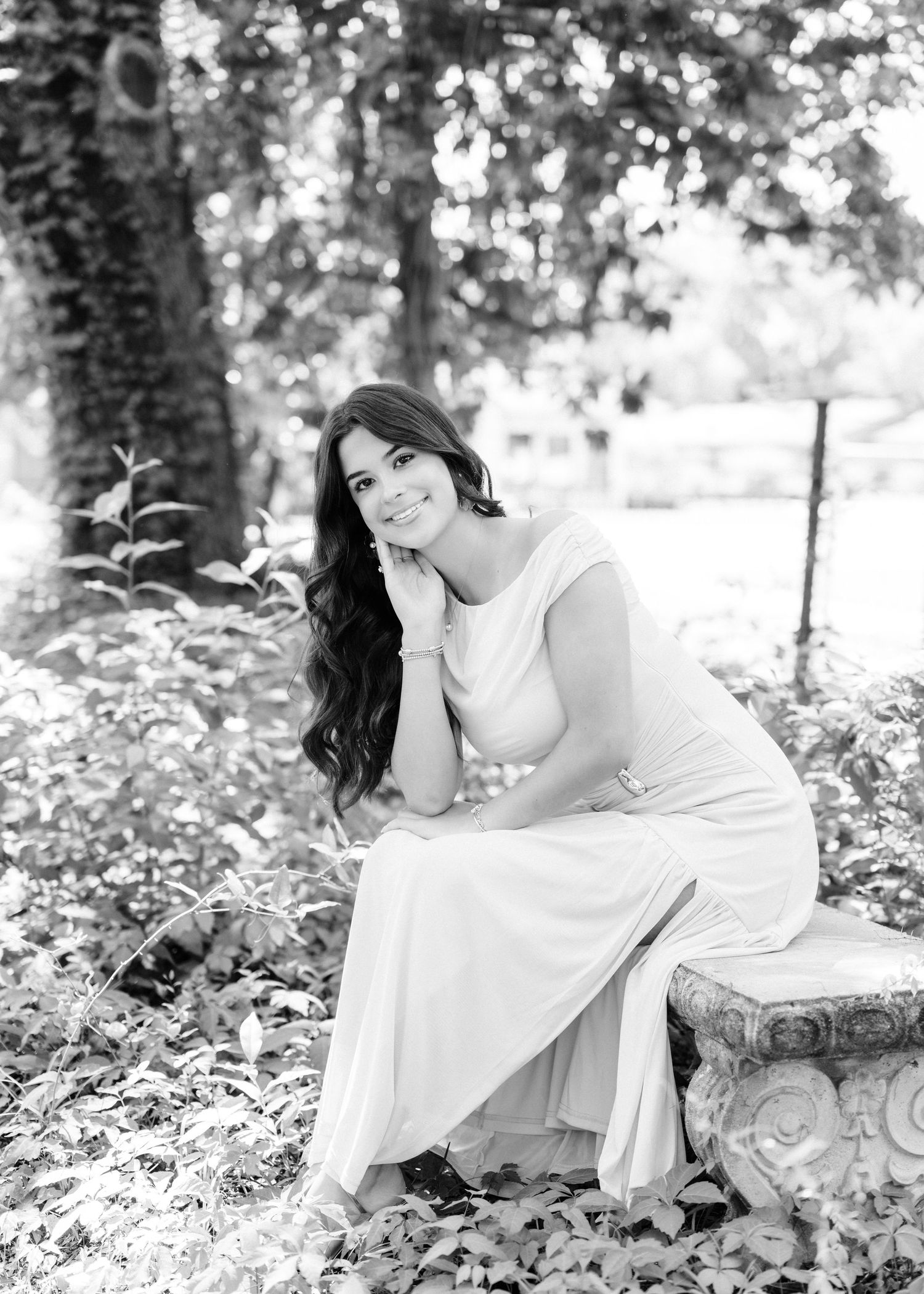 Black and white portrait of a young woman in a white dress sitting on a stone bench surrounded by lush garden foliage.