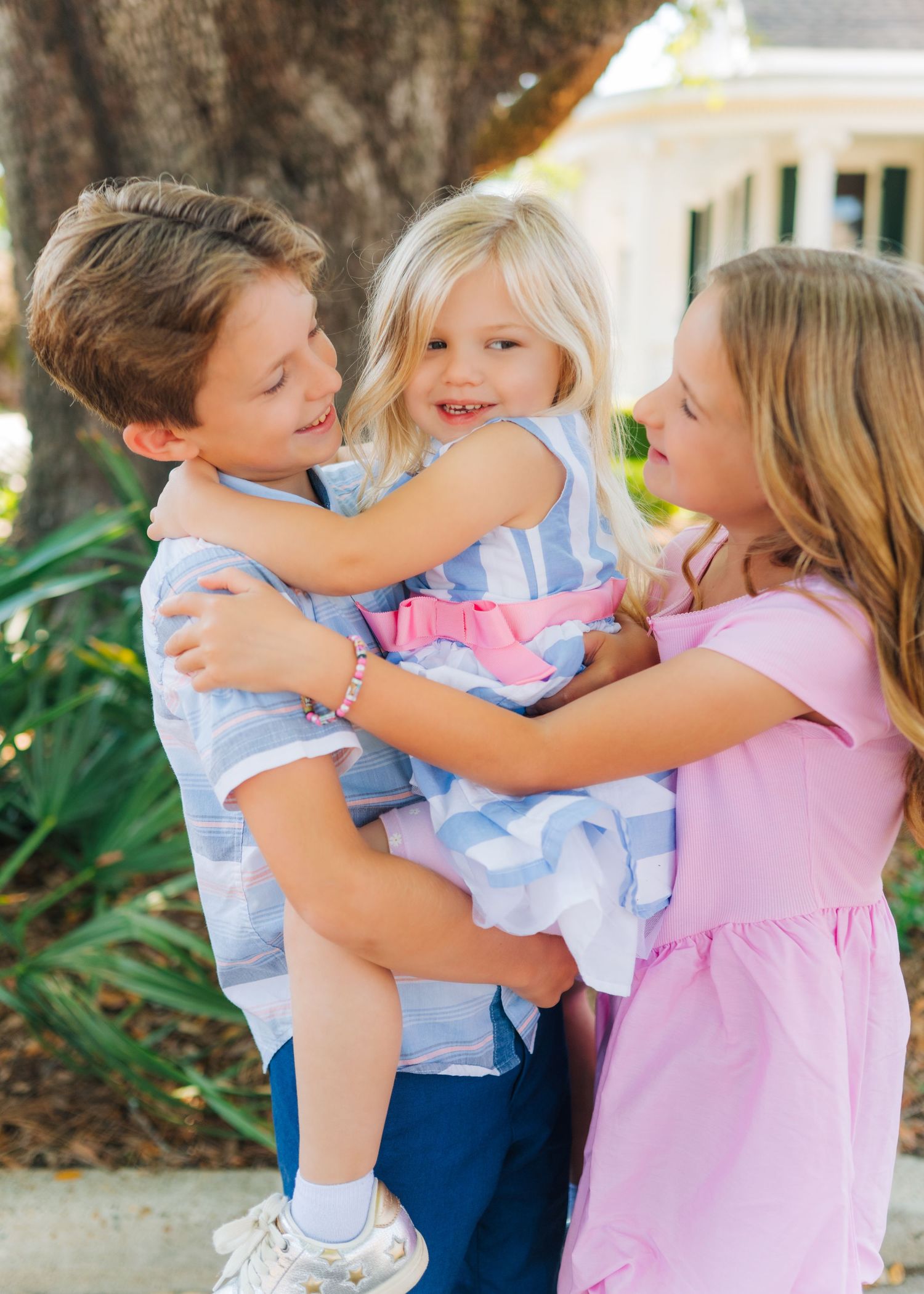 A boy holding a toddler girl while an older girl in a pink dress hugs them both outdoors near a tree.