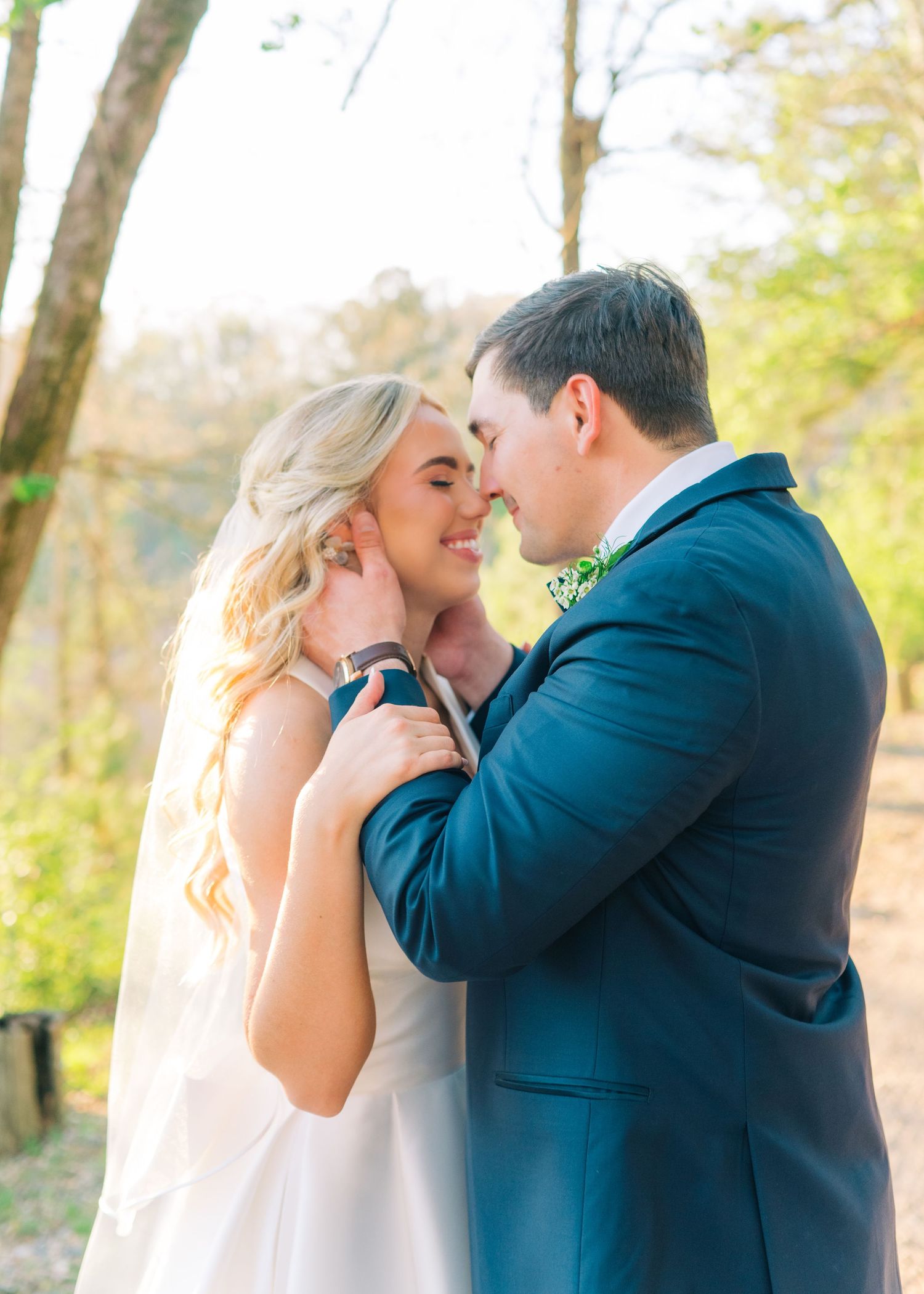 Bride and groom sharing tender moment outdoors, groom in navy suit with floral boutonniere.
