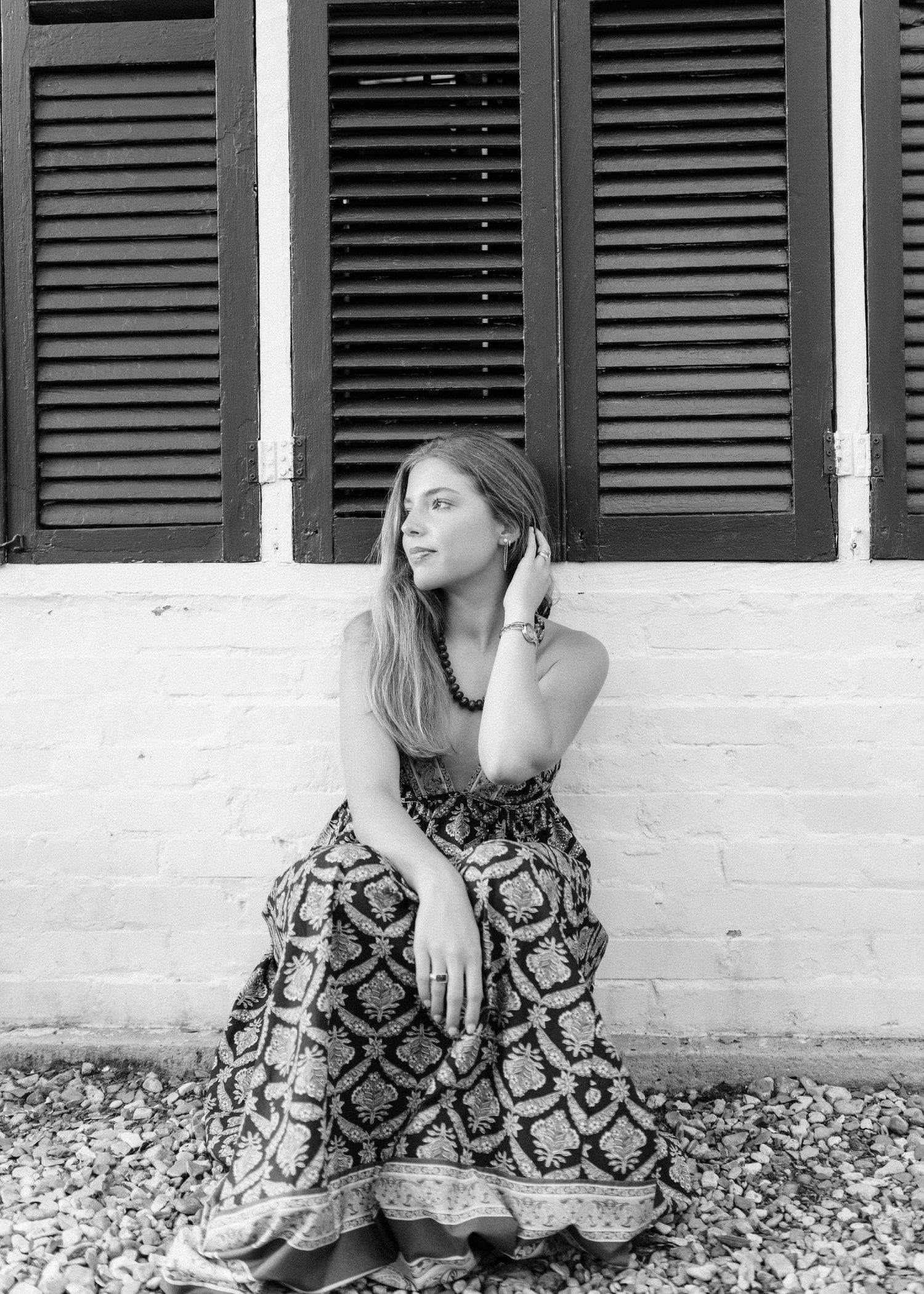 Young woman in patterned skirt sits against white brick wall with dark shutters in black and white.