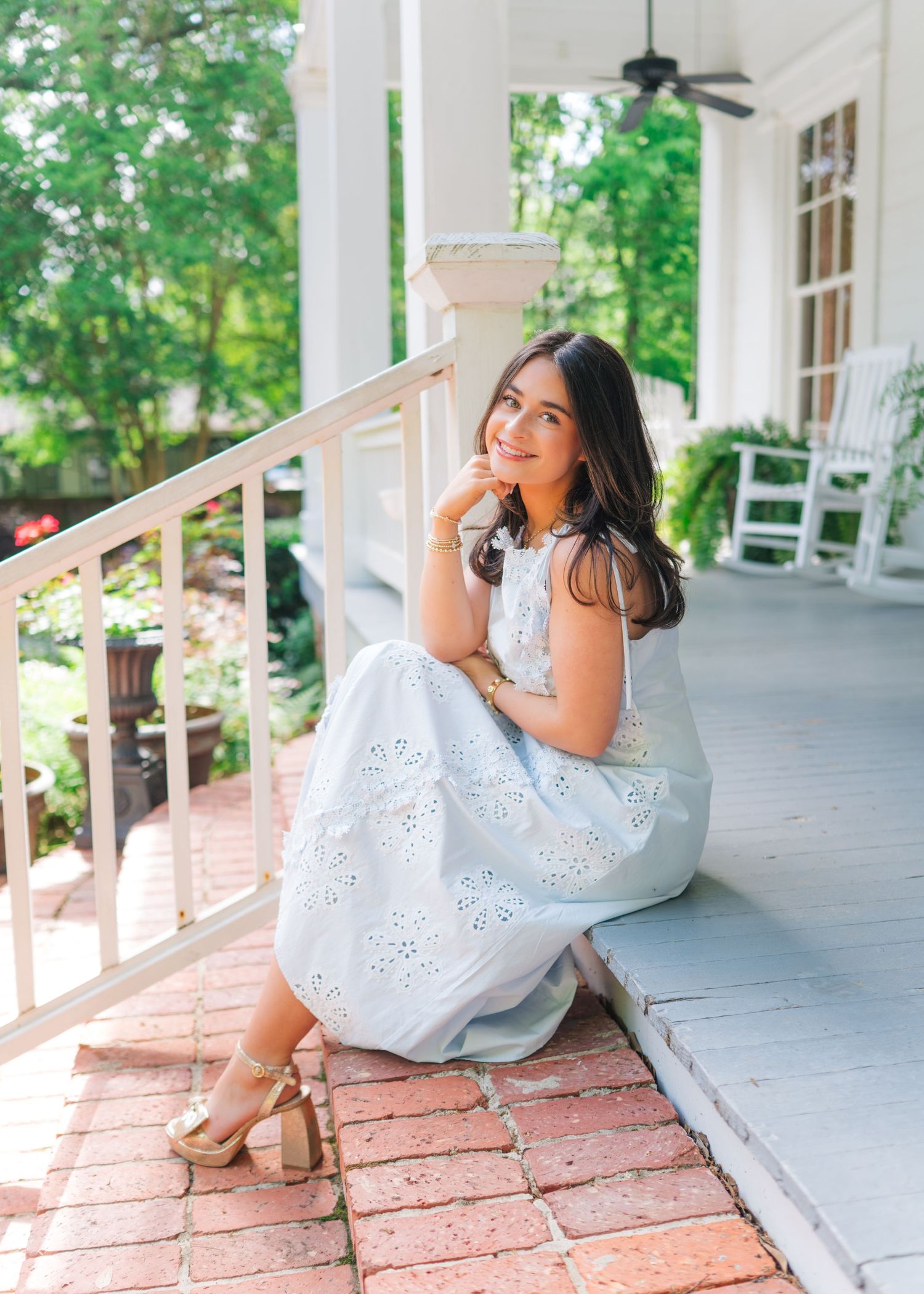 Young woman in white eyelet dress sitting on brick porch steps, smiling dreamily in lush garden setting.