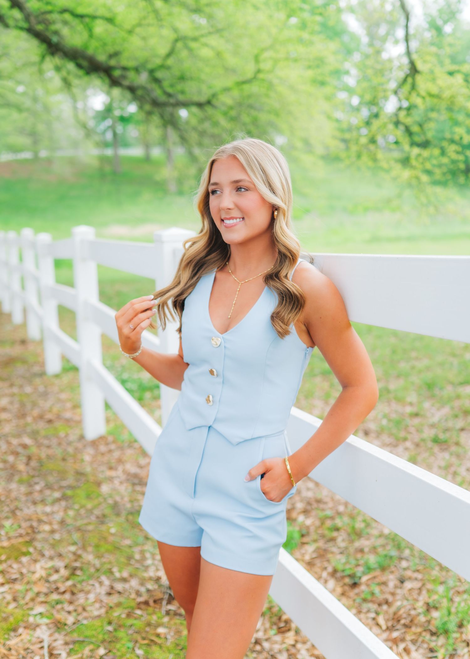 Young woman in light blue romper leaning against white fence outdoors, smiling, with lush green trees in background.