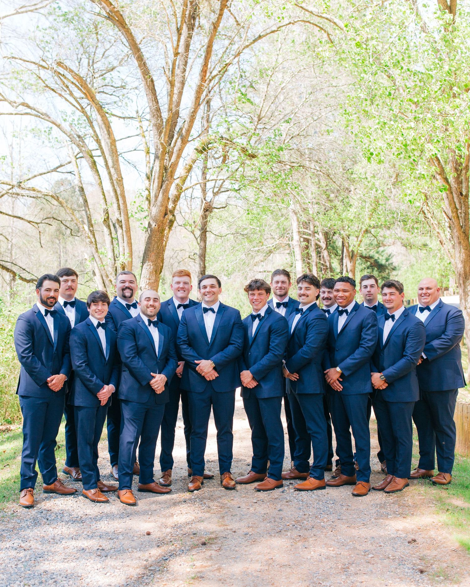 Groom and groomsmen in matching navy suits and bow ties posing outdoors under trees.