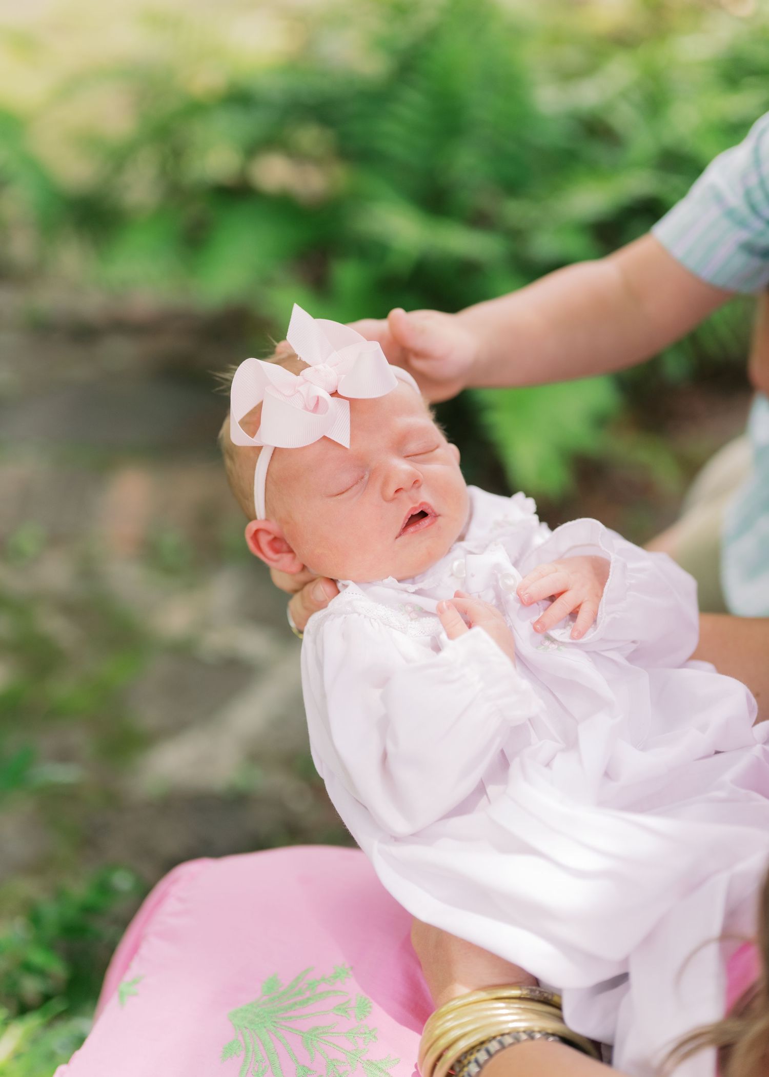 Newborn baby girl in white dress and pink bow headband sleeping peacefully outdoors while being held.