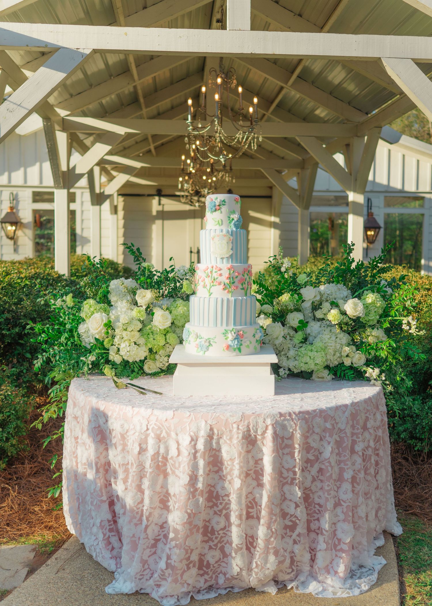 Tall white wedding cake with floral details displayed on pink lace table under a gazebo with chandelier.