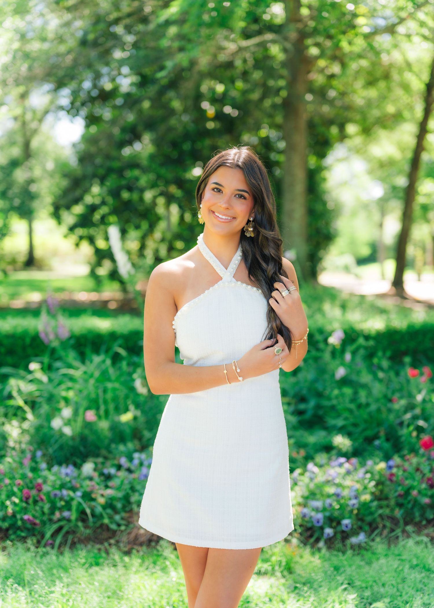 Young woman in white halter mini dress posing in lush green park with colorful flowers.