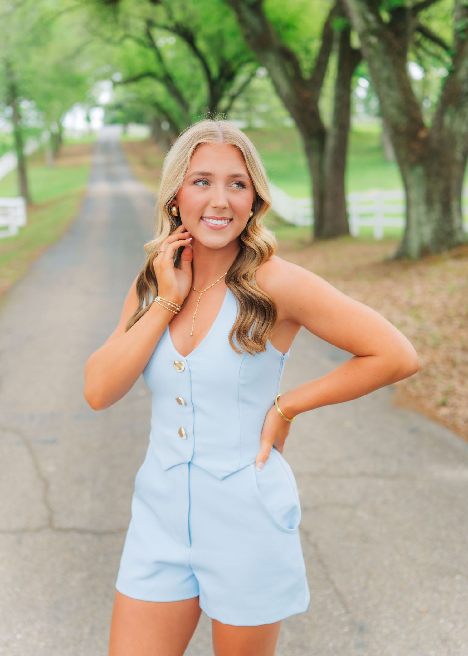 Young blonde woman in light blue romper poses on tree-lined driveway, smiling and touching her earring.