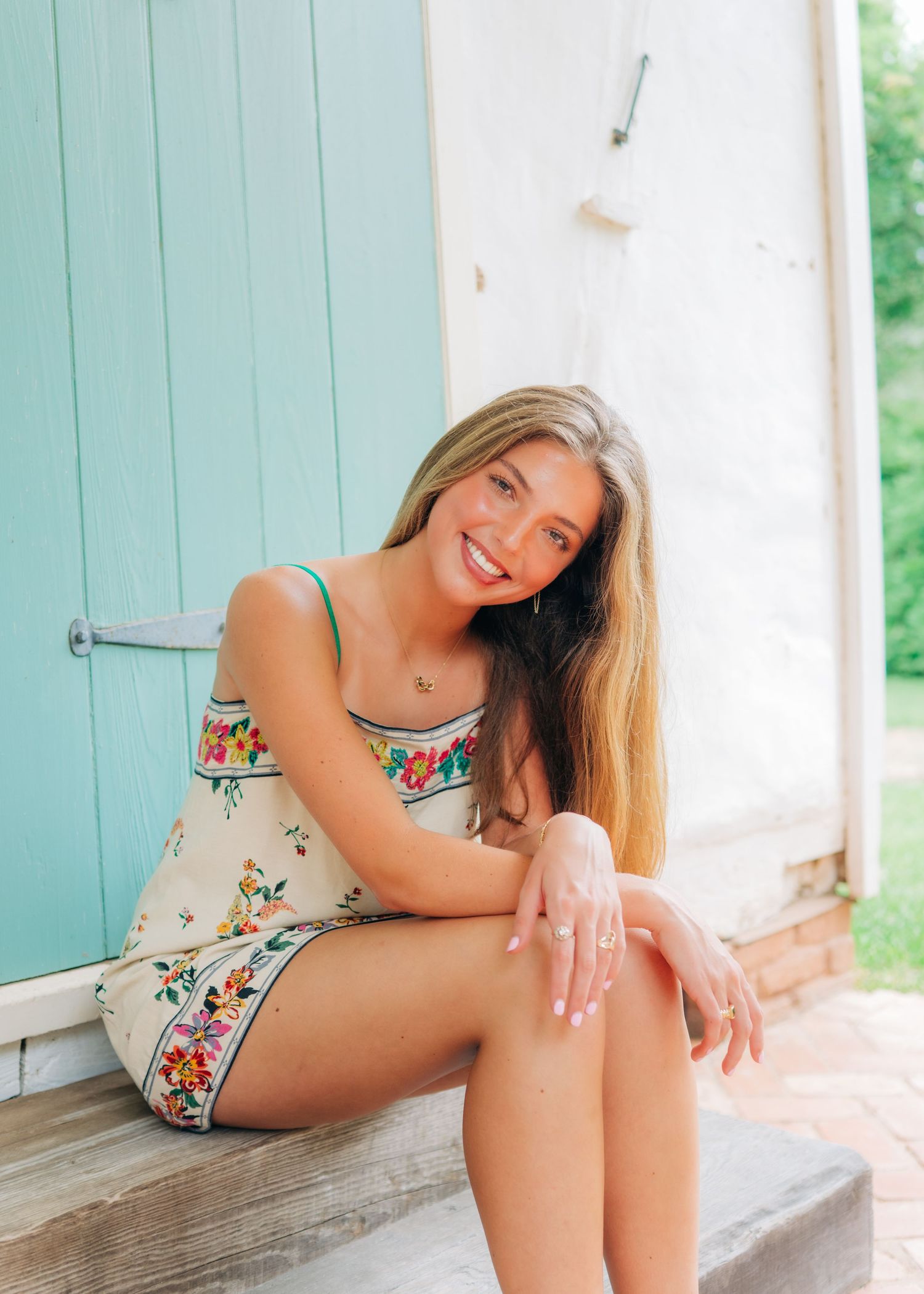 Smiling young woman in white floral dress sits on steps in front of a teal painted door.