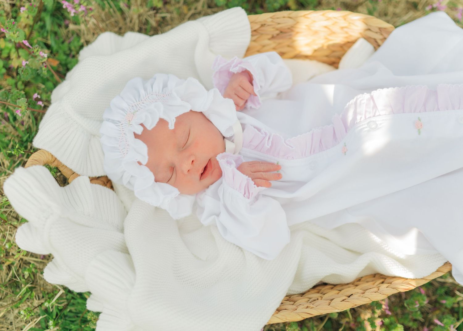 Newborn baby sleeping peacefully in a wicker basket, dressed in white outfit and bonnet on a knit blanket outdoors.
