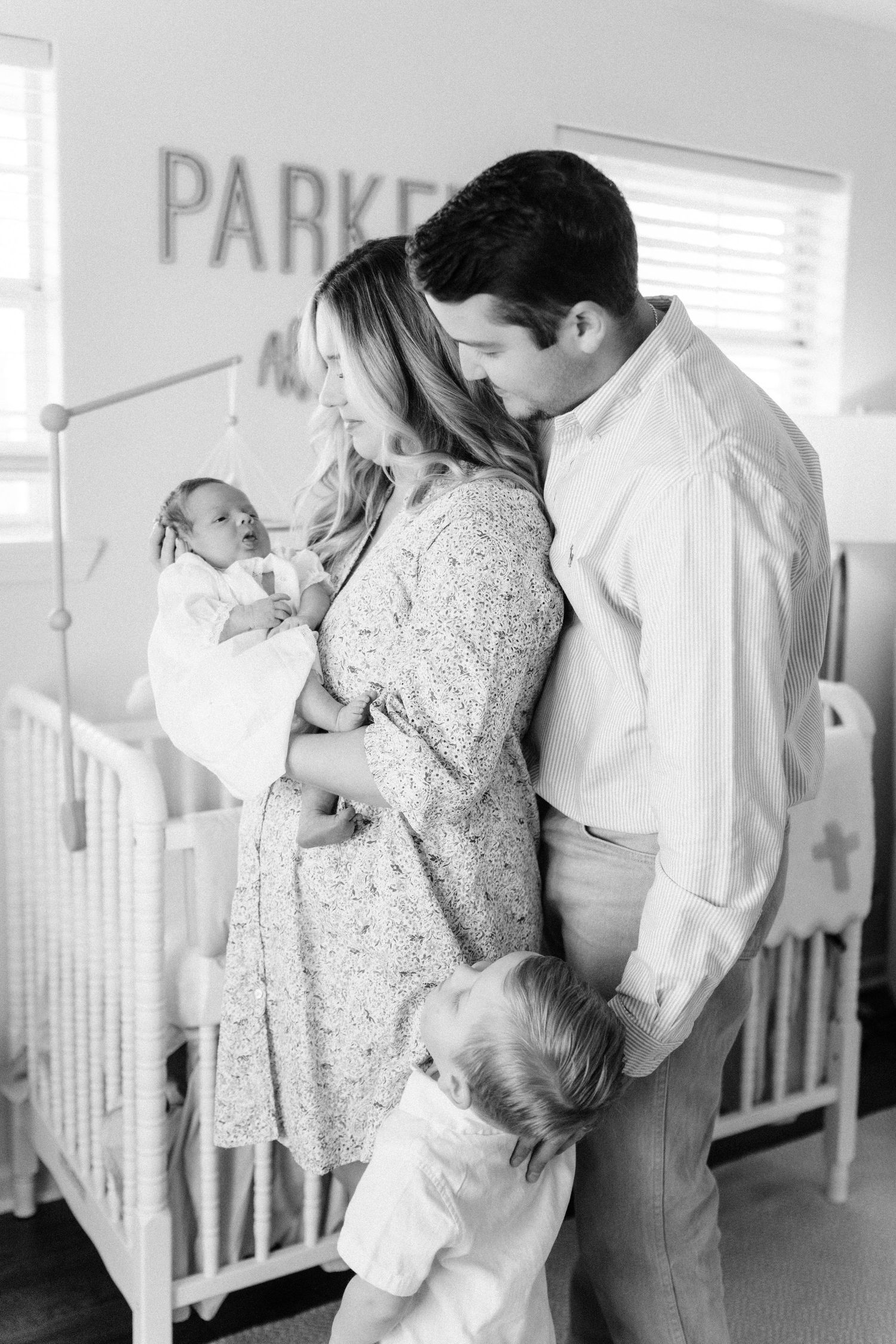 Black and white photo of parents embracing while holding newborn as toddler stands in front in nursery.