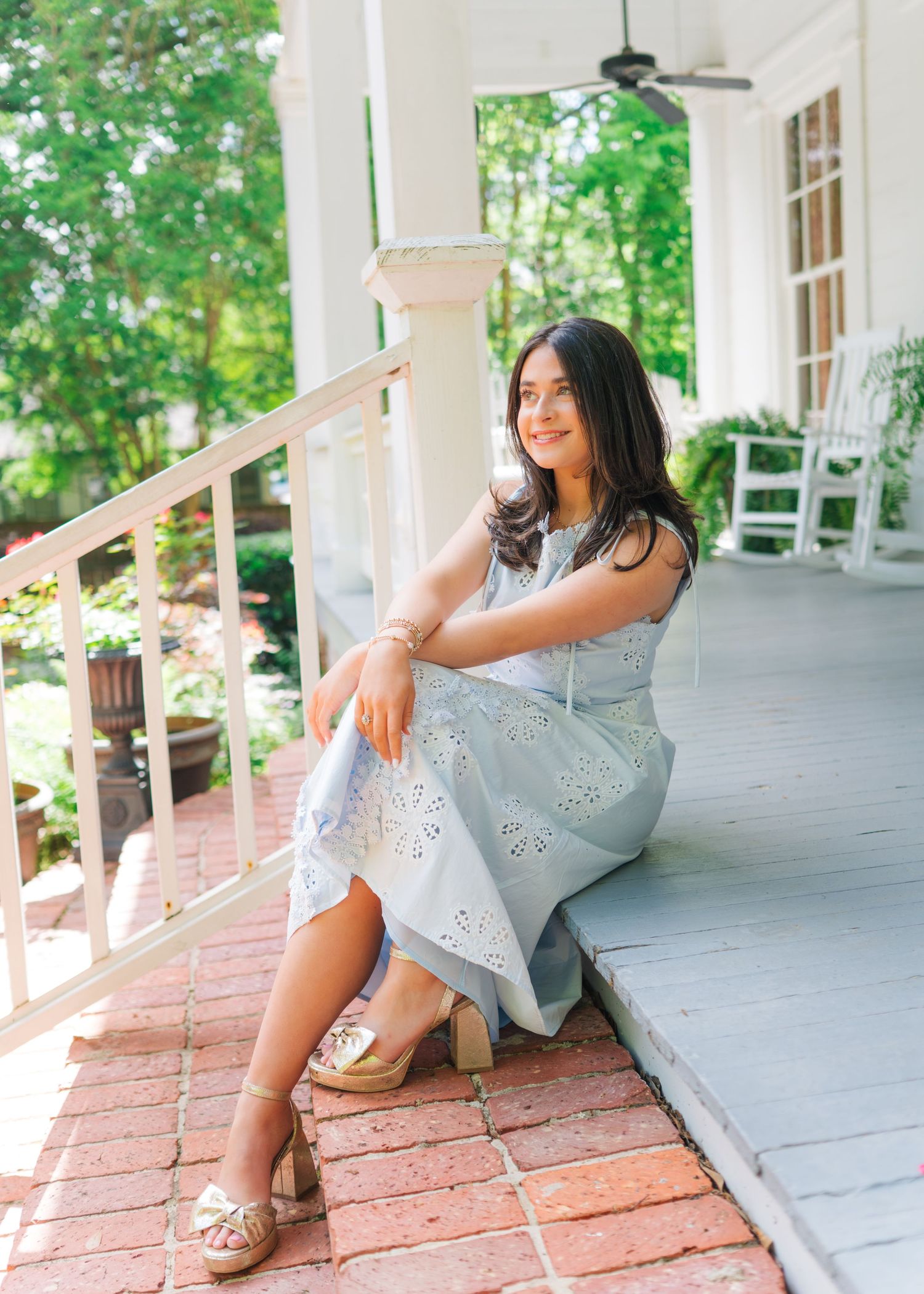 Young woman in light blue eyelet dress sitting on brick porch steps with lush greenery behind her.