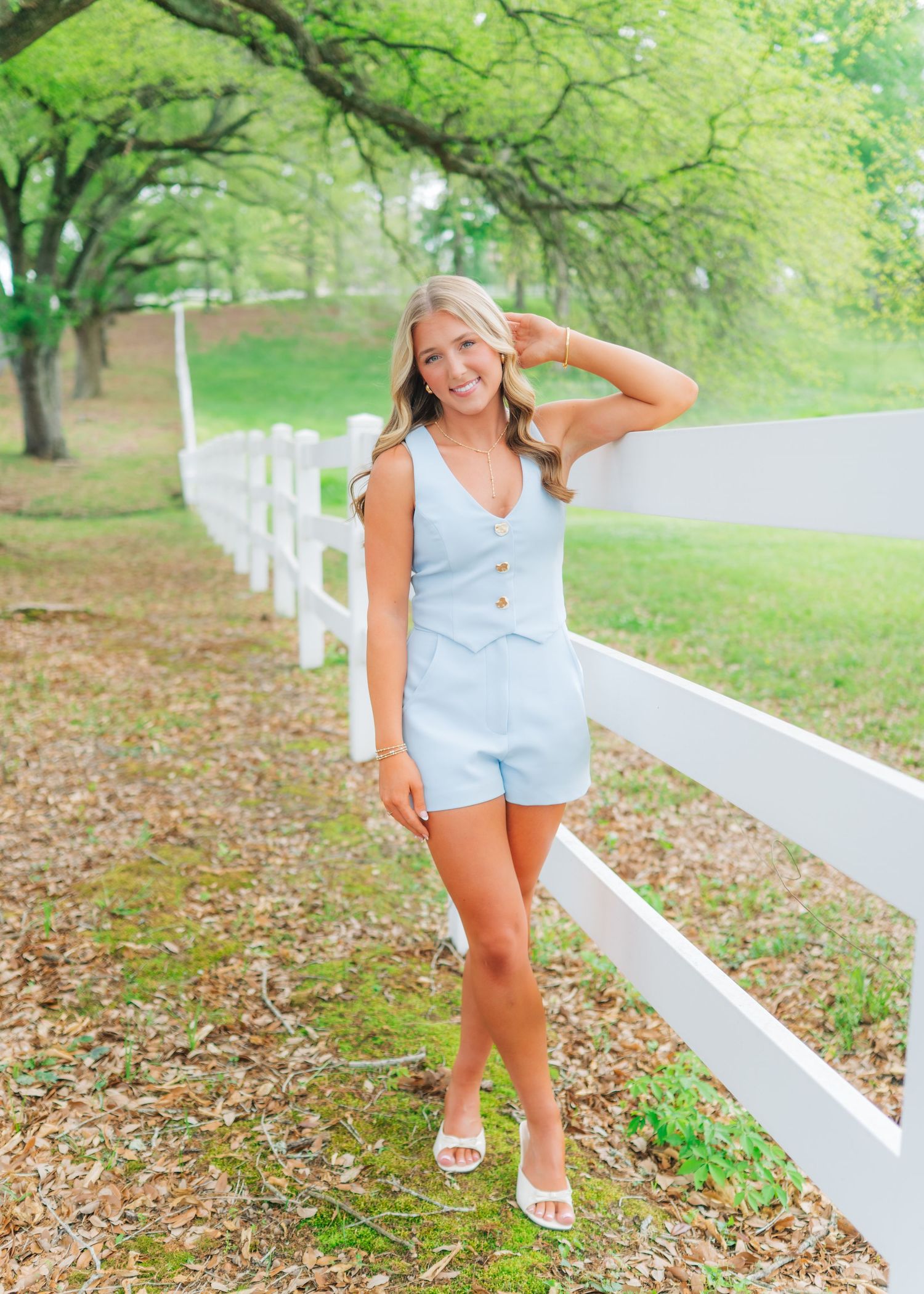 Smiling blonde teen in light blue romper poses by white fence surrounded by green trees for senior photos.