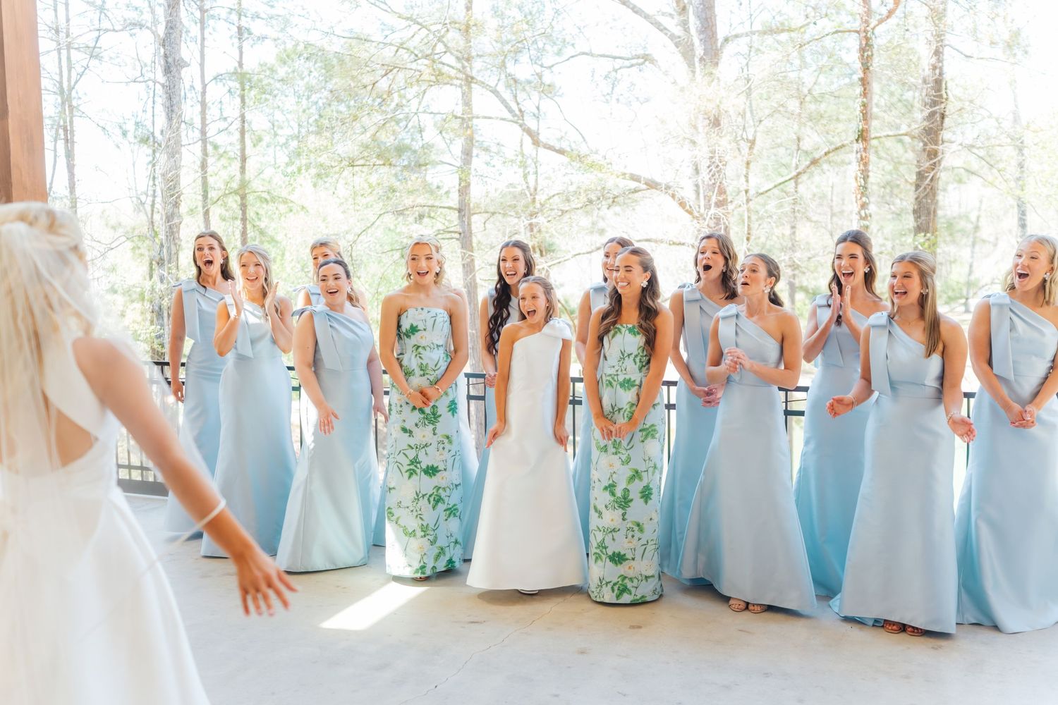 Bridesmaids in light blue gowns react with joy seeing the bride in her wedding dress at an outdoor venue.