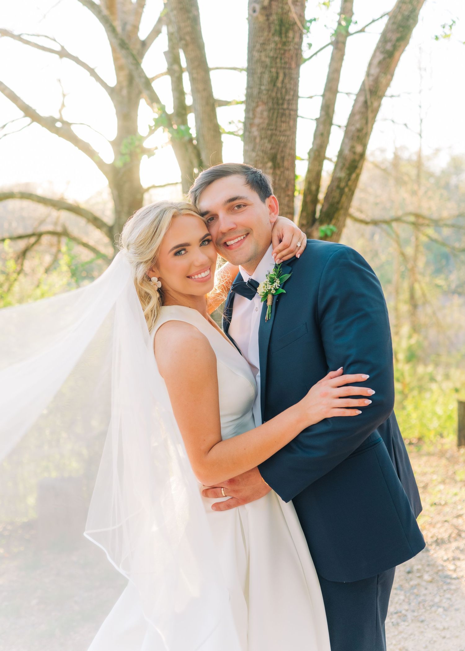 Smiling bride and groom embrace outdoors with veil flowing in breeze beneath sunlit trees.