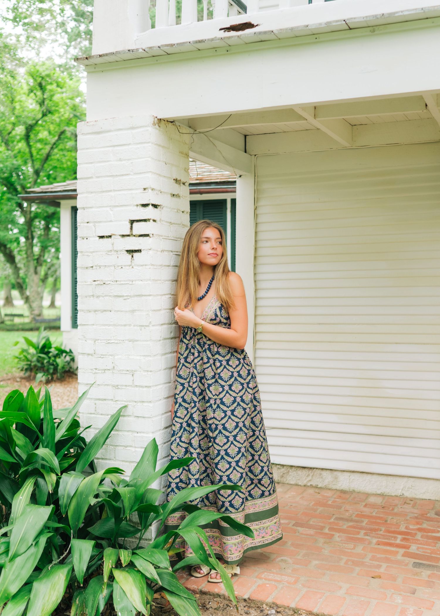 Woman in patterned maxi dress leaning against white brick building entrance.