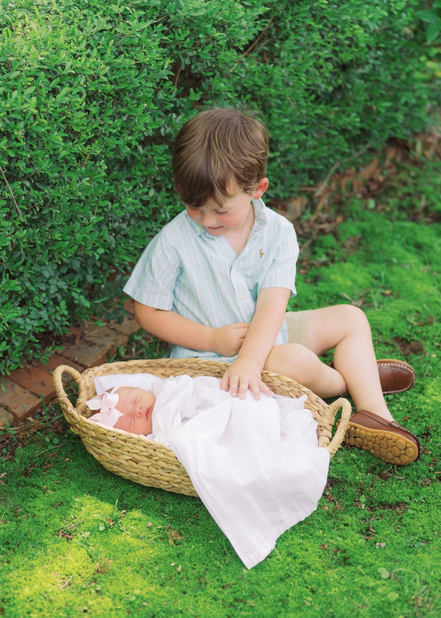 Young boy sits on green grass outdoors, lovingly watching newborn baby in a wicker basket with pink bow.