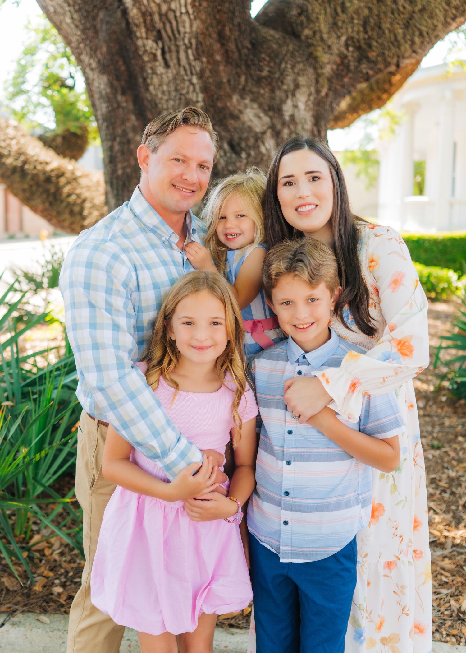 Happy family of five posing outdoors under a large oak tree, dressed in pastel spring outfits.