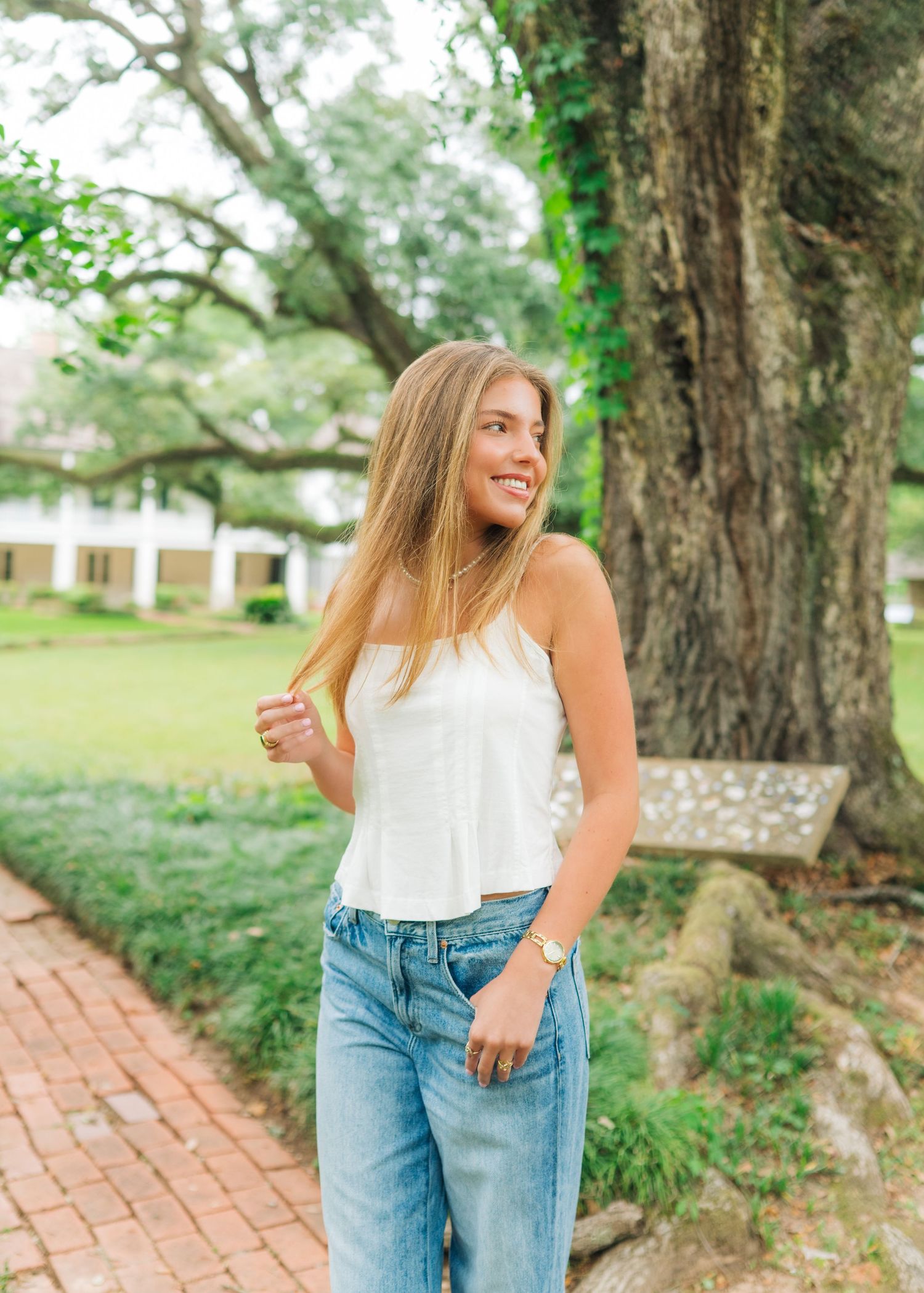 Young woman in white crop top and jeans smiling outdoors near large oak tree on brick pathway.