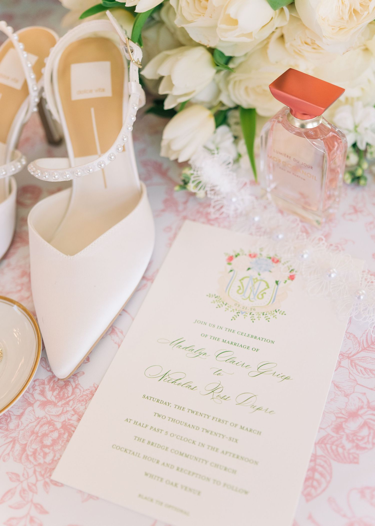 Wedding invitation with floral crest beside white heels, perfume, and white roses.