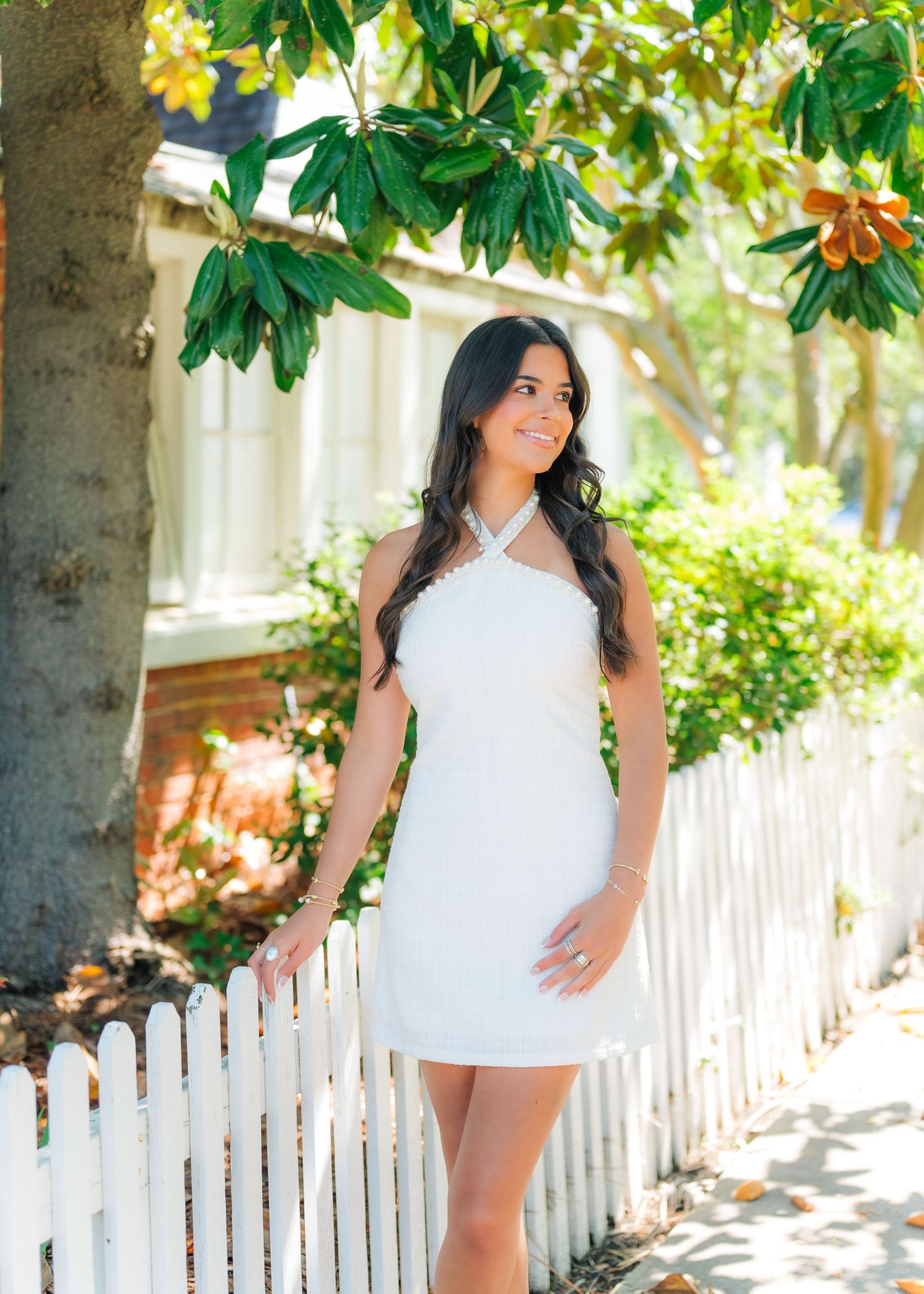 Young woman in white halter mini dress smiling outdoors by white picket fence with lush green trees.