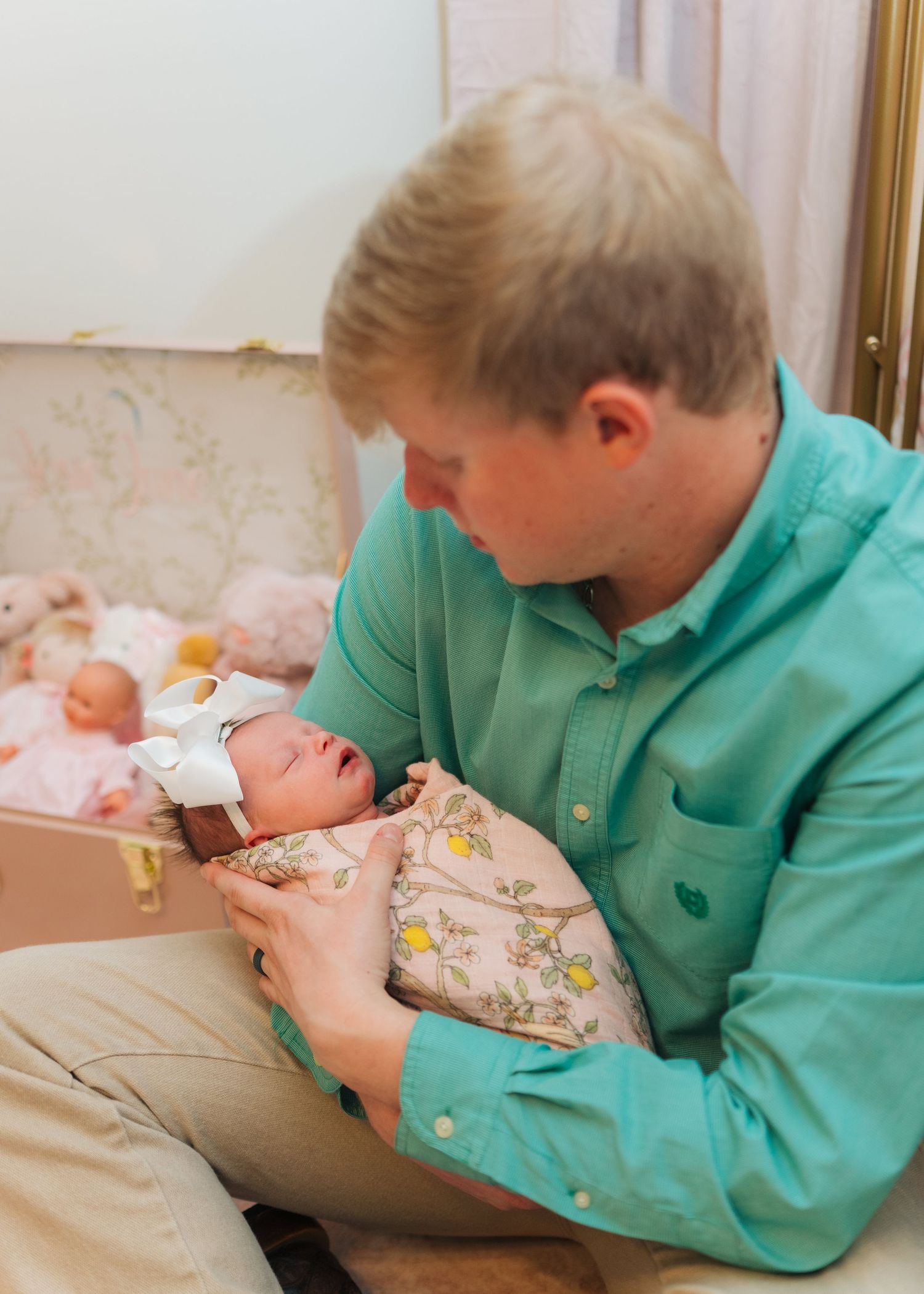 Father in teal shirt tenderly holding his swaddled newborn baby with a white bow headband indoors.