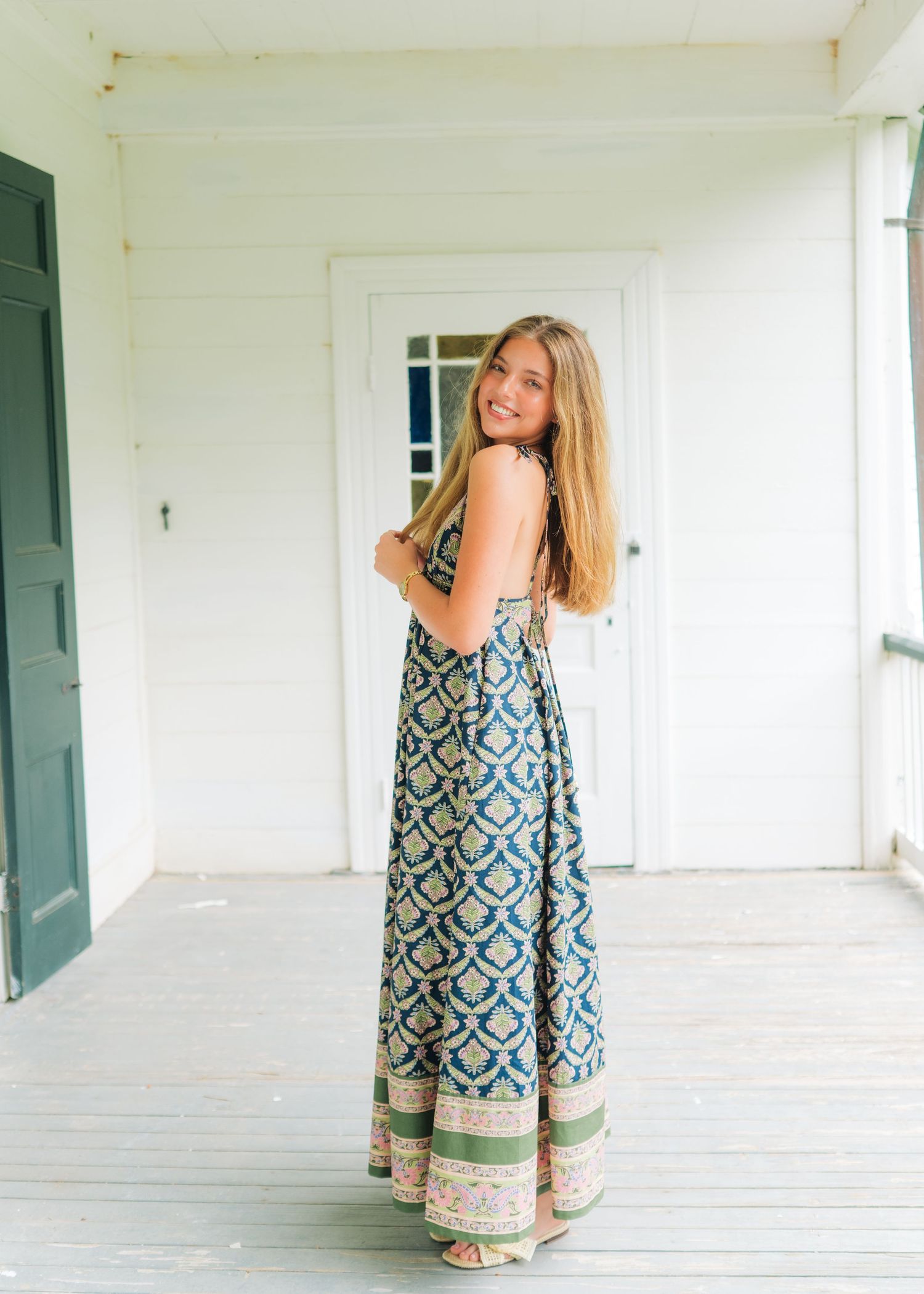 Young woman in patterned boho maxi dress smiling over shoulder on white wooden porch.