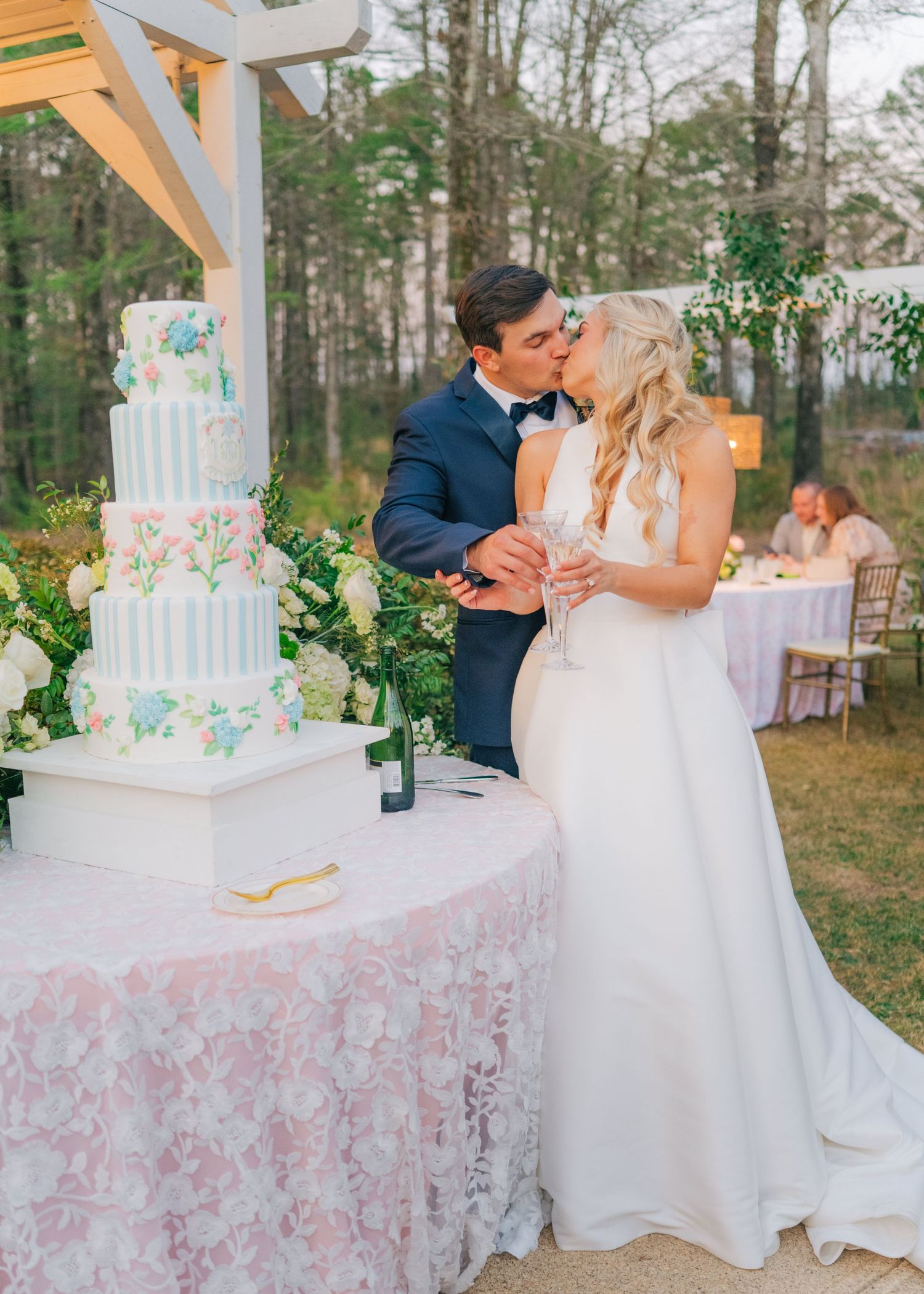 Bride and groom share a kiss beside a floral wedding cake at an outdoor garden reception.